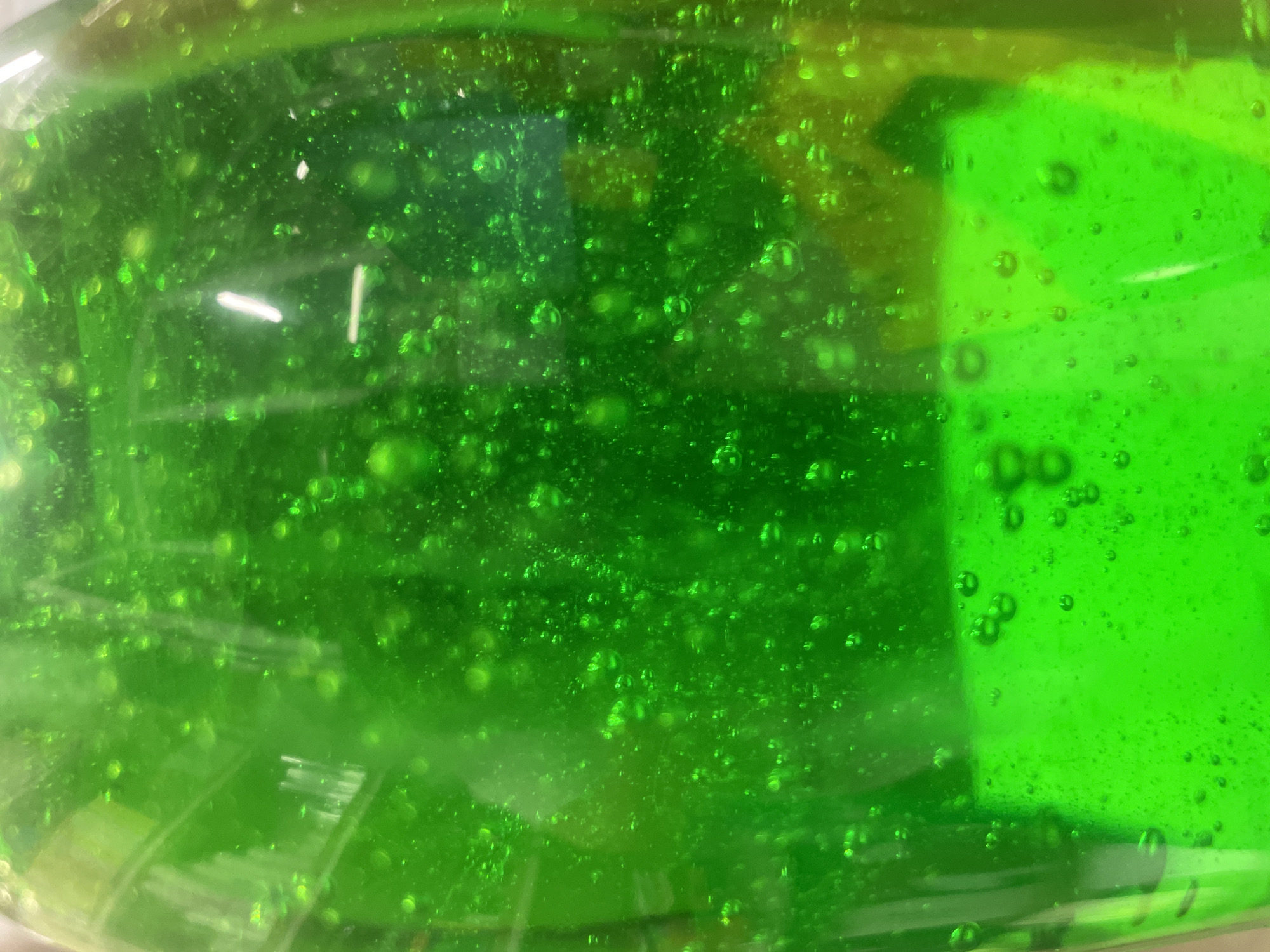 This is a close-up, eye-level, indoor shot of a vibrant green, viscous liquid, possibly hand sanitizer or a similar product, contained within a clear plastic bottle. The liquid is filled with numerous small, iridescent bubbles, some clustered together, creating a textured, almost effervescent appearance. Light reflects off the surface of the liquid and the bubbles, creating bright highlights. A distinct horizontal white line is visible in the upper right quadrant, likely a reflection from a light source. The lower portion of the image shows a flatter, more uniform layer of the green liquid with a slightly different texture, suggesting a separation or a change in the liquid's density. The background is a blurred, out-of-focus green hue, consistent with the liquid's color. The scene is devoid of any people or other identifiable objects, focusing solely on the visual properties of the liquid itself. There is no discernible connection to the city of Терволово, Russia, or any urban environment.