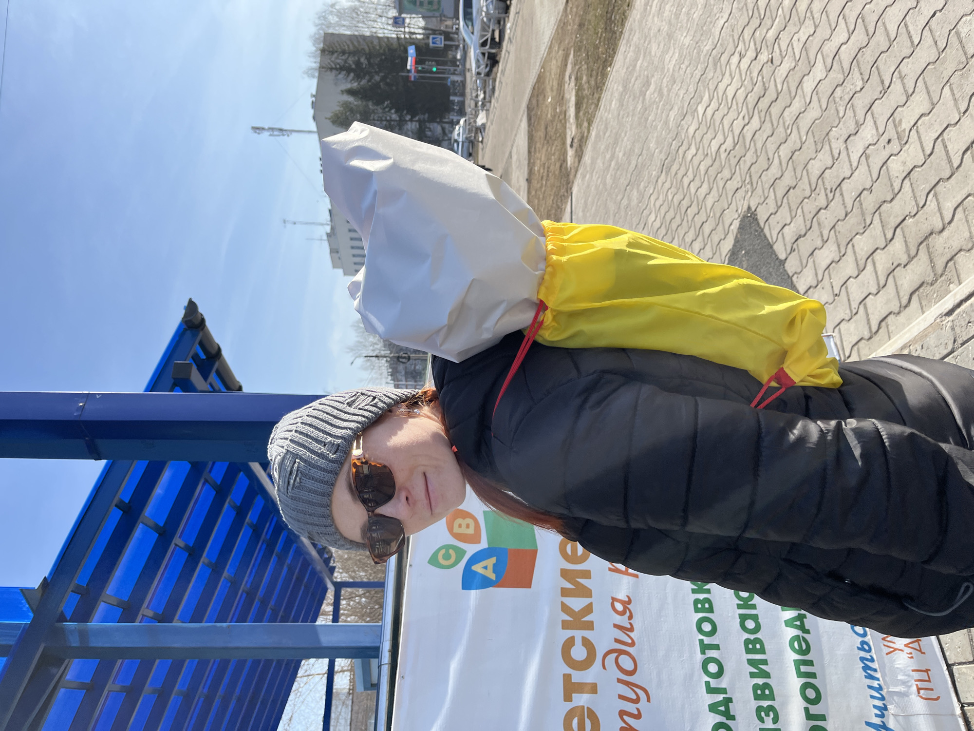A woman stands outdoors on a sunny day in Chaikovsky, Russia. She wears a grey knit hat and sunglasses, and a black puffer jacket. A large white and yellow bag sits behind her head, attached to her shoulders with red drawstrings. To her left is a blue structure, likely a bus stop. Beside her is a white sign with colorful lettering and Cyrillic text, advertising детские студии (children's studios). The background includes a street, buildings, and trees, indicating an urban setting. The ground is paved with interlocking bricks.