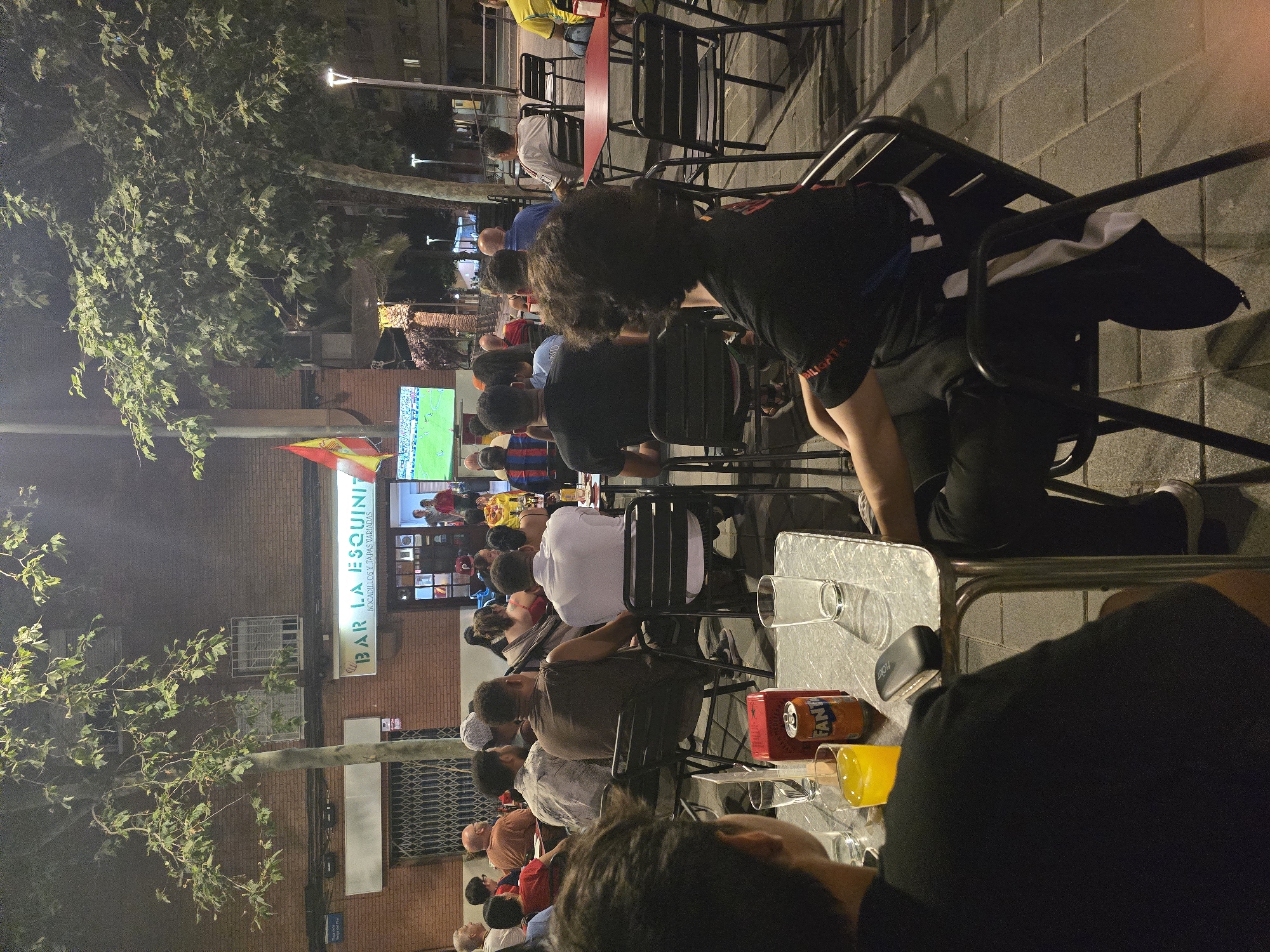 A group of people are seated at tables outside a bar named "BAR LA ESQUINITA", with a Spanish flag visible. They are watching a soccer match on a television screen. The bar has a brick facade and metal security gates. The scene is illuminated by ambient lighting and streetlights. A Fanta can and glasses of orange juice are on a table in the foreground. The image was taken at night.