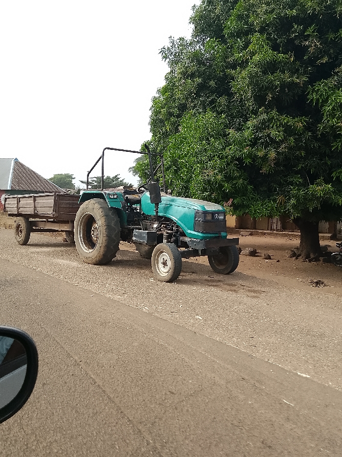 An teal-colored tractor with a brown, two-axle wooden trailer is positioned on the dirt shoulder adjacent to a paved road in Jibam, Nigeria. The tractor's large rear tires are soiled, and a black roll bar is visible above the operator's area. A large, densely leafed green tree occupies the right side of the frame, with bare earth and small rocks at its base. In the background, to the left, a building with a dark-colored corrugated metal roof is partially visible. The sky is bright white and overcast. A portion of a dark vehicle's side mirror is visible in the lower left corner, indicating the vantage point of the photographer. No individuals are visible.
