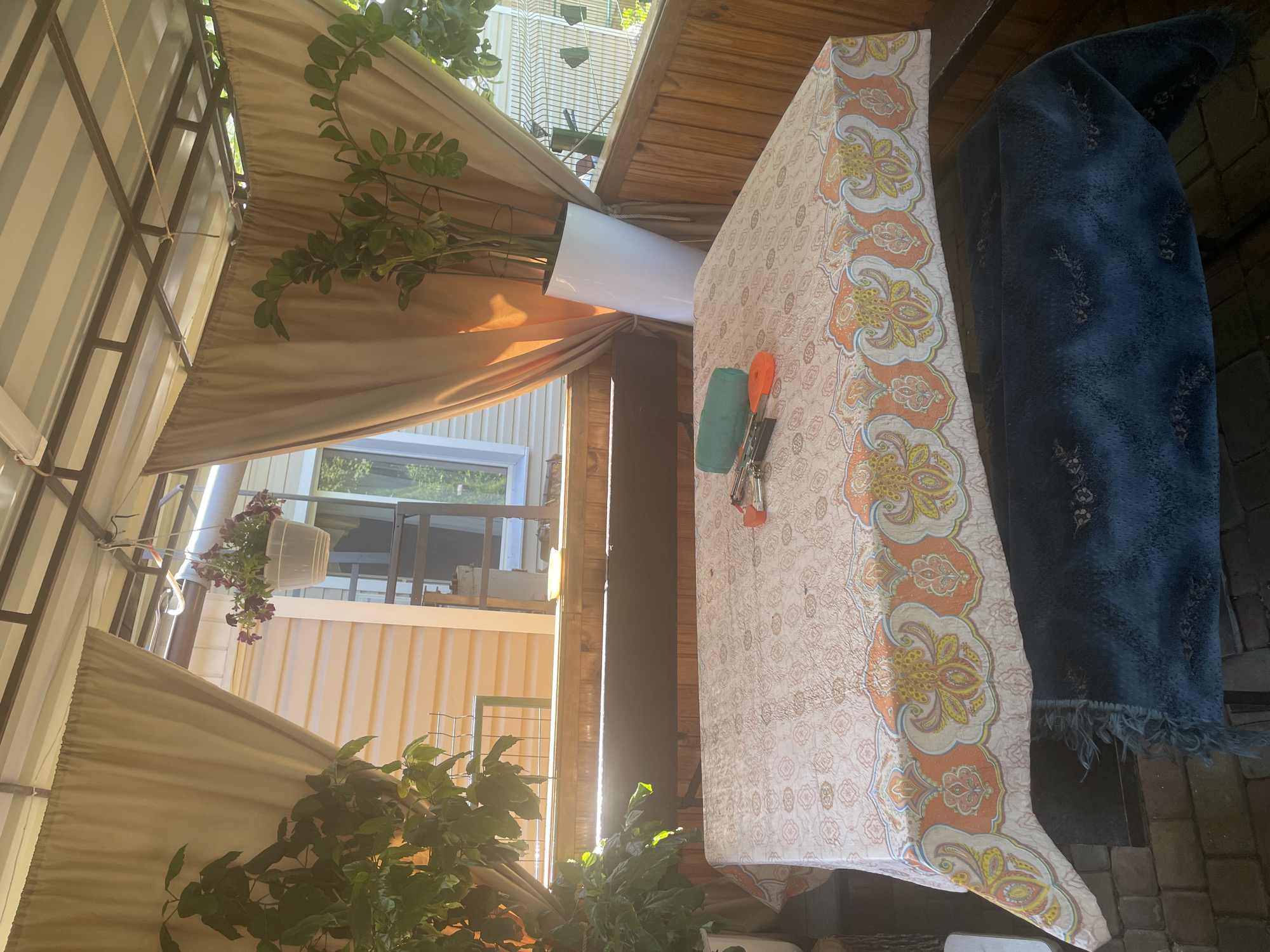 The image depicts an outdoor covered patio area, likely in the city of Vysokyi, Ukraine. The scene is set with a table covered by a tablecloth featuring a patterned design of orange and yellow paisley-like motifs. On the table, several objects are visible, including a green cylindrical item, a pair of scissors, and what appears to be a keyring with multiple keys.
To the left of the table, a large potted plant with dark green leaves is situated, casting shadows on the tablecloth. A second plant, taller and also green, stands in a white cylindrical pot to the right of the table. In the background, a house with pale yellow siding is visible, with windows and a portion of a second story. The patio itself is constructed with wooden walls and a metal roof structure, supported by beams. Beige curtains hang from the roof, framing the view of the house. Beneath the table, a dark blue textured fabric, possibly a rug or blanket, is draped over a bench. The ground beneath the patio appears to be paved with bricks. The lighting suggests it is daytime, with sunlight filtering through the plants and curtains.