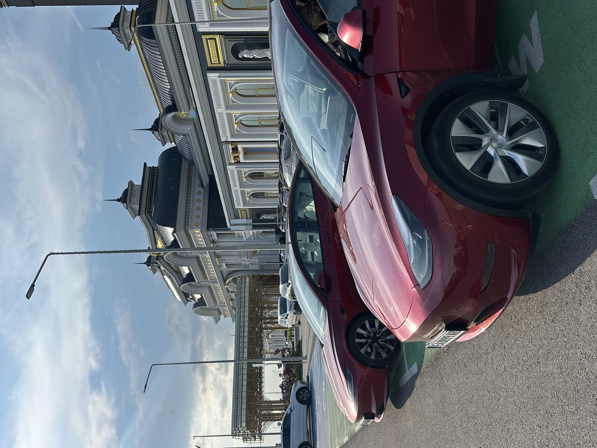 This detailed image captures an outdoor scene at the Kapitan Andreevo border crossing in Kapitan Andreevo, Bulgaria, featuring two red Tesla electric vehicles parked in the foreground, set against a backdrop of an ornate, large building under a partly cloudy sky.
In the immediate foreground, a red Tesla Model Y is partially visible on the right, showing its front passenger side, including the wheel, fender, and part of the hood and windshield. A person with dark hair and glasses is visible inside this Tesla, looking towards the right side of the frame through an open passenger window. Immediately behind and to the left of the Model Y, a red Tesla Model 3 is parked. Its front bumper displays a Bulgarian license plate, partially legible as "E 8899 AB." Both Teslas are parked on asphalt, within parking spaces marked with green paint.
In the middle ground, a grand, classically-styled building dominates the scene. It features multiple dark, domed roofs adorned with golden accents and spire-like finials. The building's facade is characterized by white and gray sections, further embellished with gold trim, ornate columns, arched windows, and decorative niches housing white statues, one of which is clearly visible on the right side of the facade. To the far left in the background, a modern, light-colored canopy structure with intricate metalwork is visible, extending over what appears to be another roadway or pedestrian area.
Several other vehicles are parked further back near the building, including a white SUV to the far left and various other cars indistinctly parked in the distance. The sky above is partly cloudy, with patches of blue sky and white clouds, and the lighting suggests either morning or late afternoon, with shadows extending from the vehicles and streetlights. Two tall streetlights are visible on the left side of the frame, and another is prominent in the upper center, partially obscuring the building.