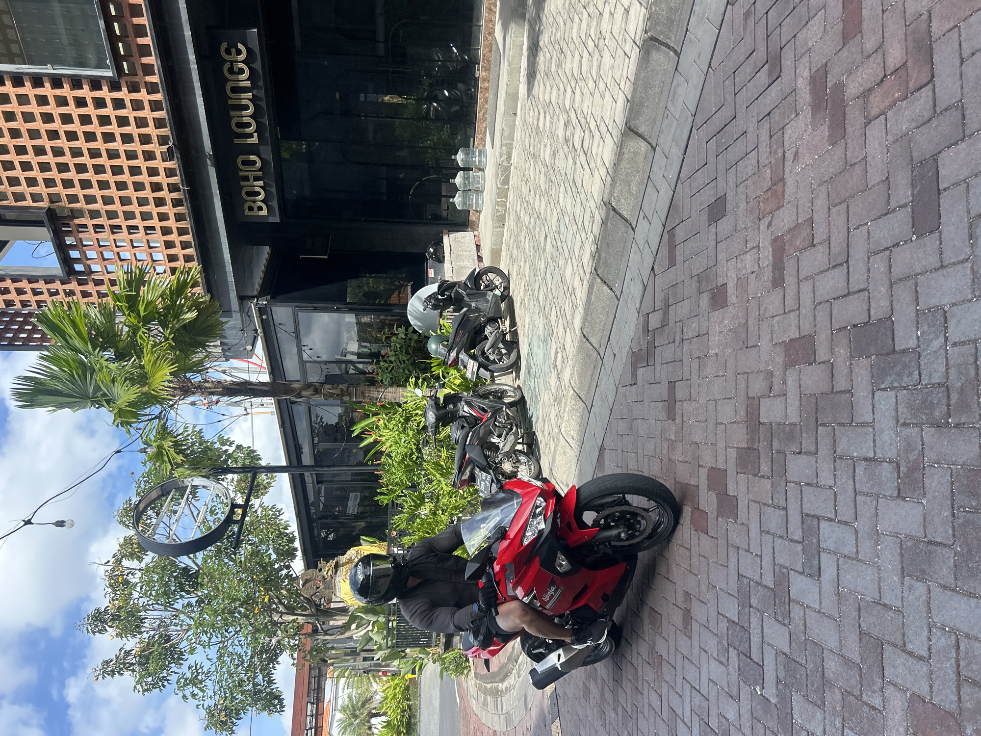 A man wearing a black shirt and black helmet is riding a red motorcycle on a brick-paved street in front of a building with a sign that reads "BOHO LOUNGE". The building has a distinctive orange brick facade with square openings. To the left of the building, there are trees and a large circular metal art installation. Two black motorcycles are parked in front of the building, and several large water cooler jugs are stacked near the entrance. The sky is blue with scattered clouds. The scene appears to be in a commercial area of North Kuta, Indonesia.
