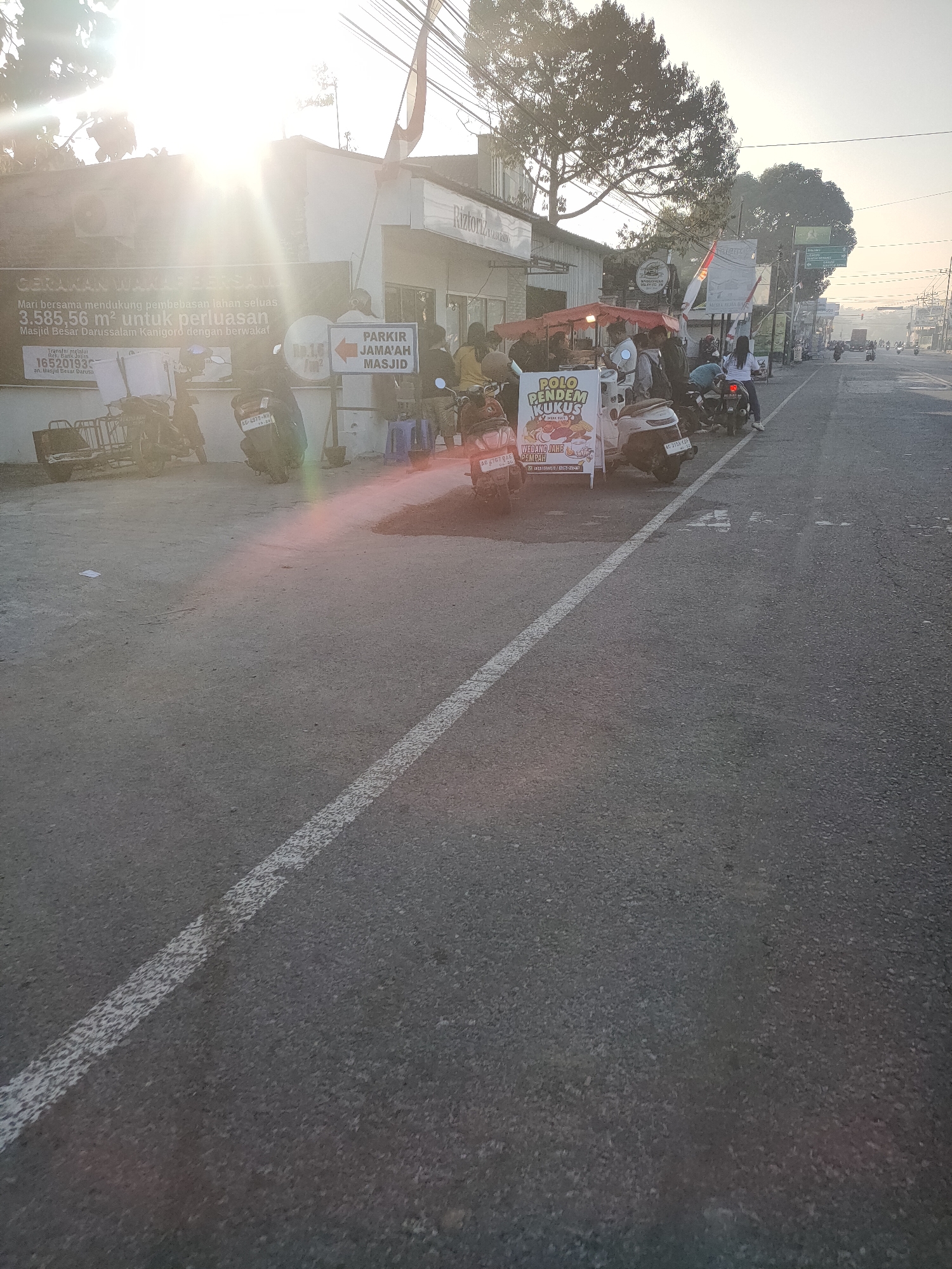 In Kanigoro, Indonesia, a street scene unfolds with a bright sun glaring from the left, casting lens flares across the image. A building with a sign that reads "Riztoriz" is visible on the left, alongside a poster advertising "GERAKAN WAKAF BERSAMA Mari bersama mendukung pembebasan lahan seluas 3.585,56 m² untuk perluasan Masjid Besar Darussalam Kanigoro dengan berwakaf." Several parked motorcycles are present, including one with a cart. A food stall selling "POLO PENDEM KUKUS" and "WEDANG JAHE REMPAH" is set up with a red awning, attracting a crowd of people gathered around it. A sign indicating "PARKIR JAMA'AH MASJID" (Mosque Congregation Parking) is also visible. The street is paved and lined with a white dashed line. In the background, more vehicles are seen on the road, with buildings and trees lining the street. The overall atmosphere suggests a busy commercial area on a sunny day.