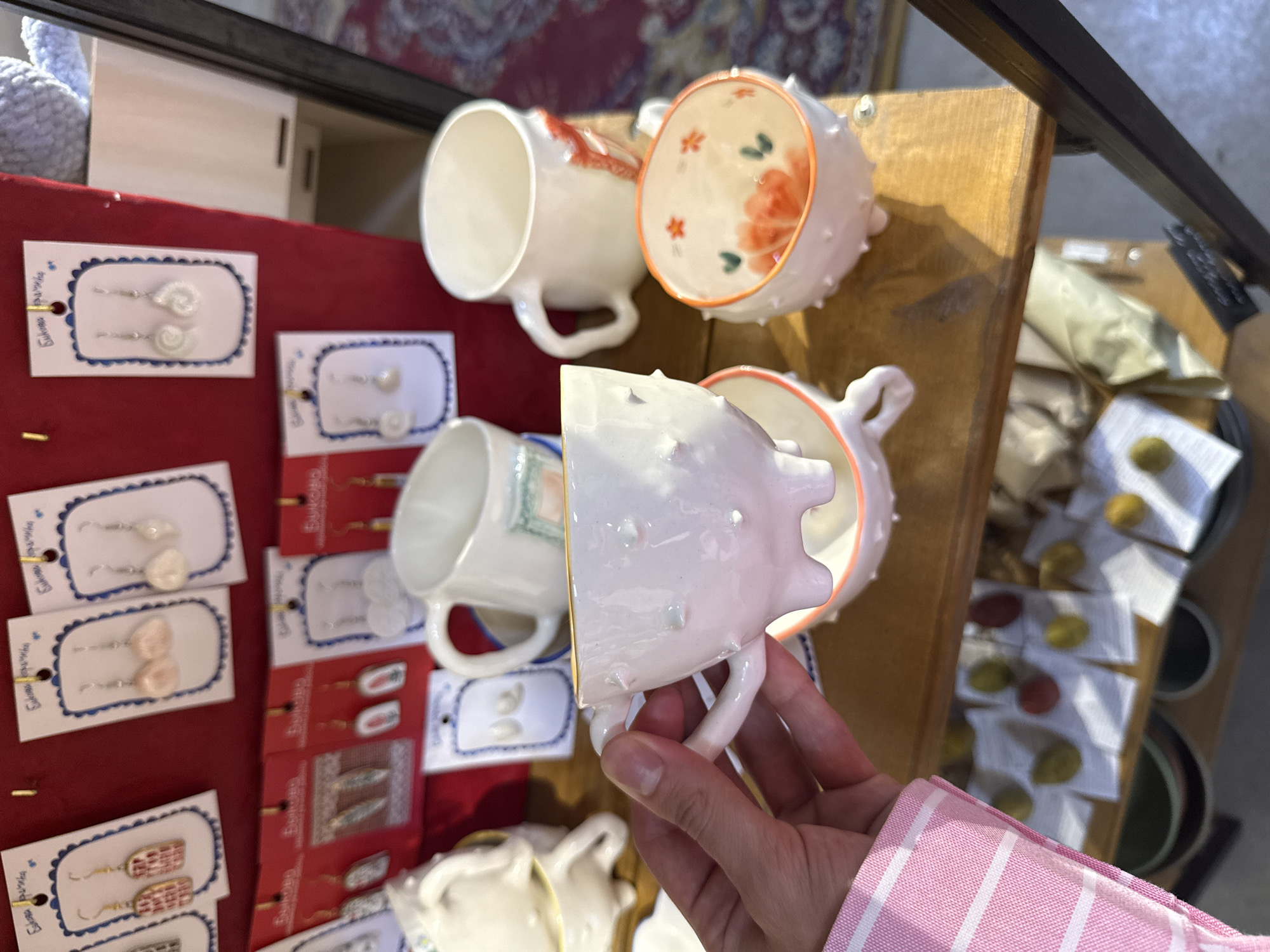 A person's hand, wearing a pink and white striped garment, holds a white ceramic mug with three small, rounded feet. The mug is textured with small, raised bumps and has a golden rim. Behind it, another white ceramic mug with a handle sits on a wooden shelf. To the right of this mug is a smaller, white ceramic bowl also resting on its feet. This bowl has an orange rim and is decorated with painted orange flowers and red star-like shapes inside.
Above the mugs and bowl, a red display board is visible, showcasing various handcrafted earrings mounted on white cards with blue borders. These earrings appear to be made of beads, shells, and other small decorative elements.
Below the shelf with the mugs and bowl, another wooden shelf holds several small, individually wrapped items, some of which appear to be fruits or similar objects, laid out on white paper. The overall scene suggests a market stall or a small shop displaying handcrafted goods, potentially in Saint Petersburg, Russia, given the context. The lighting indicates it is daytime, with sunlight casting shadows.