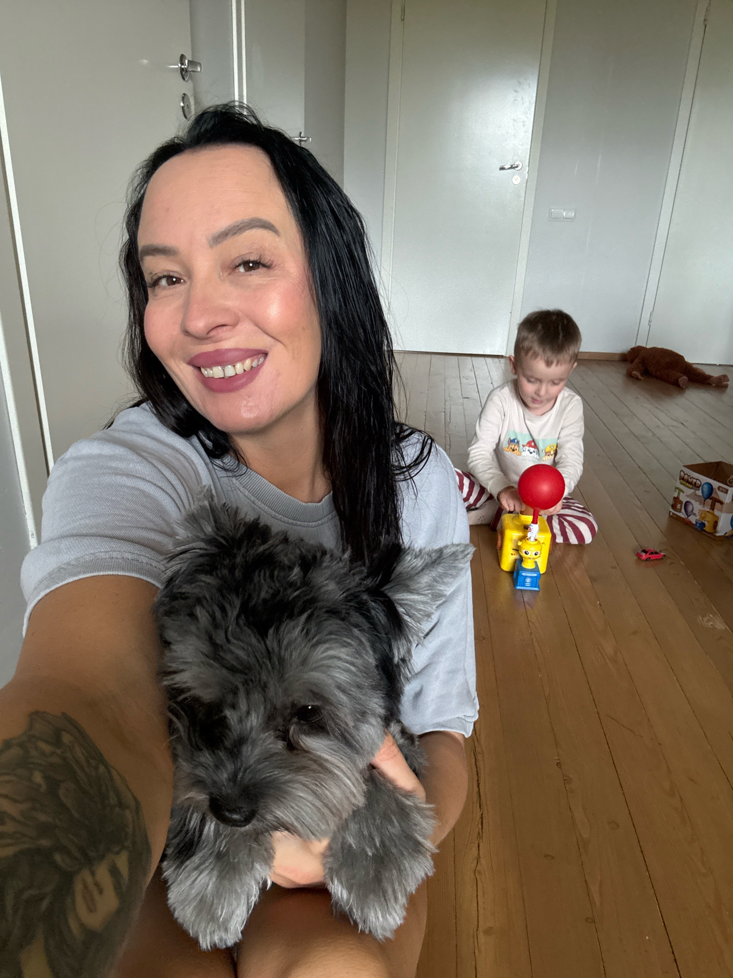In Chaikovsky, Russia, a woman with long black hair and makeup smiles for the camera, holding a gray schnauzer puppy. She wears a light gray t-shirt and has a tattoo on her left arm. In the background, a young boy in a white shirt and striped pants plays with a red balloon on a yellow toy. The room has light-colored walls with doors and a wooden floor. A brown stuffed animal and a toy car are visible on the floor.