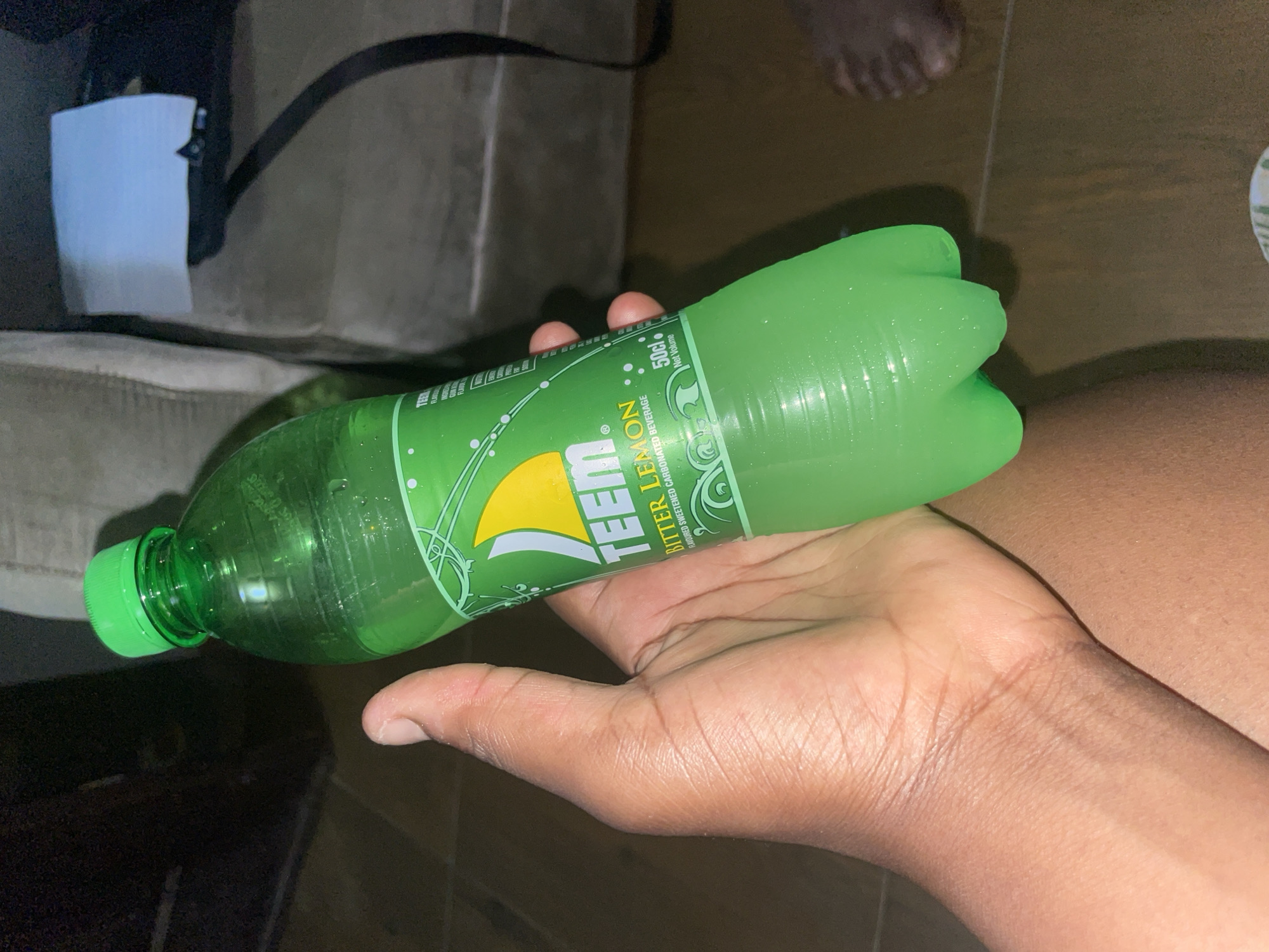 A green bottle of Teem Bitter Lemon soda is being held in a person's hand. The location appears to be indoors. Additional details include a partial view of a person's foot in the background, a white box, and a black strap. There is no information about the location being in Kurunduma, Nigeria.