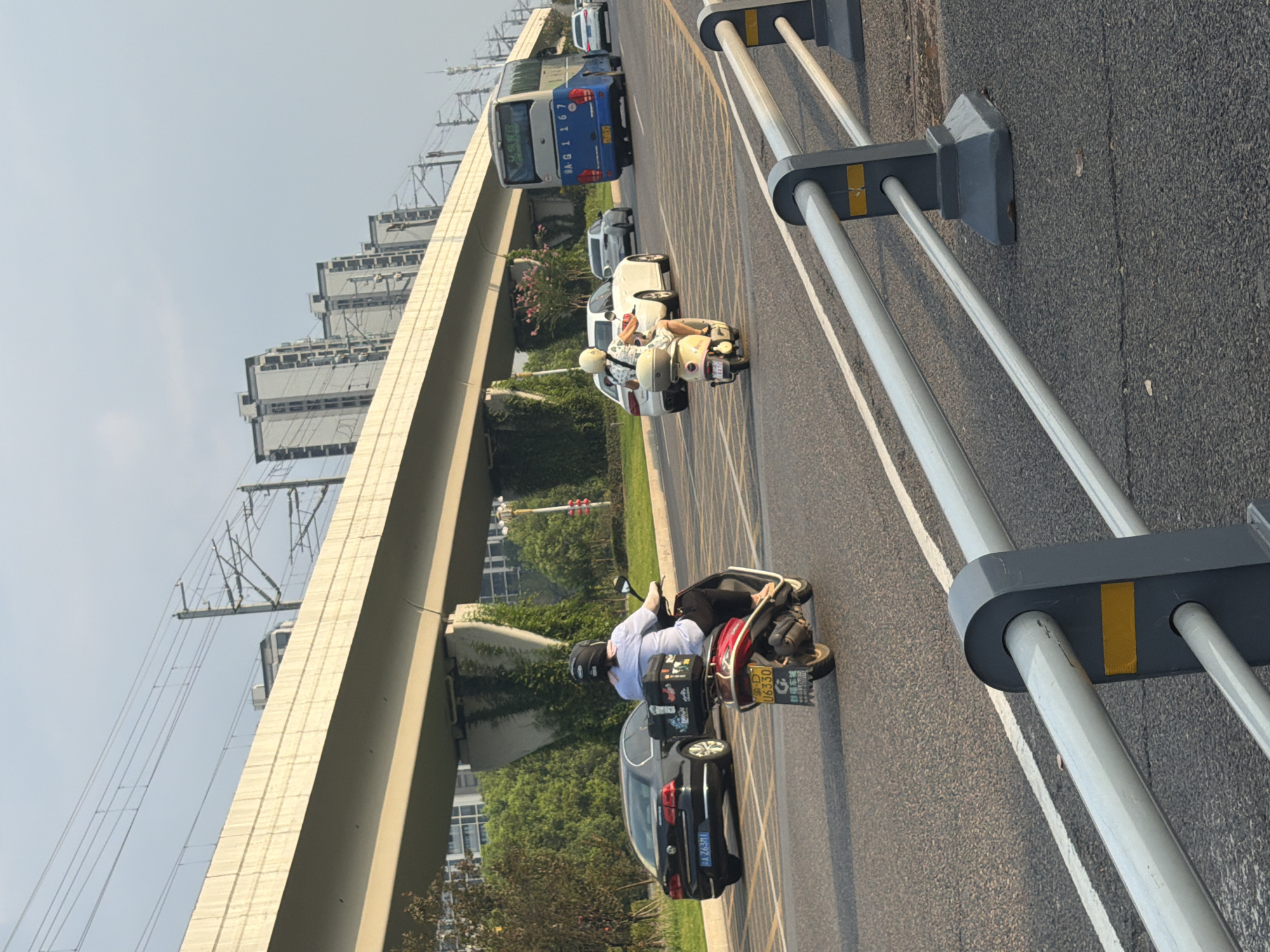 This image depicts a street scene in Jiangjin, China, featuring a multi-lane road with a pedestrian overpass or bridge overhead. The overpass supports railway tracks and overhead power lines. Buildings and greenery are visible in the background.
In the foreground, a red scooter is being ridden by a person wearing a helmet and a light blue shirt. The scooter has a black storage box mounted on the rear, with various stickers or labels on it. A black car is parked or moving on the left side of the road. Further down the road, a light yellow scooter is being ridden by a person wearing a white outfit and a helmet. Several other cars and a blue bus are also present on the road.
The road surface is asphalt, with white lane markings. A grey metal guardrail with yellow reflective markers runs along the right side of the road, separating lanes or providing a barrier. The sky is clear with some haze, and the lighting suggests it is either early morning or late afternoon. The overall environment is urban, with elements of transportation infrastructure, vehicles, and residential or commercial buildings.