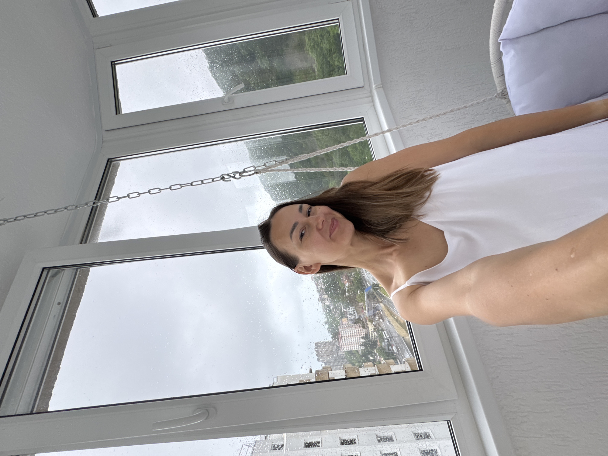A woman with long brown hair is taking a selfie in an enclosed balcony with large windows. She is wearing a white sleeveless top. The windows show a cityscape with buildings and trees, and it is raining, with water droplets visible on the glass. The overall mood is casual and relaxed, with the woman smiling at the camera. The scene appears to be indoors, likely an apartment or hotel room, with a white textured wall visible on the right. A white cushioned chair or swing is partially visible on the upper right, attached to the ceiling by ropes and chains. The lighting suggests it is daytime, but the overcast, rainy weather makes it appear dim.