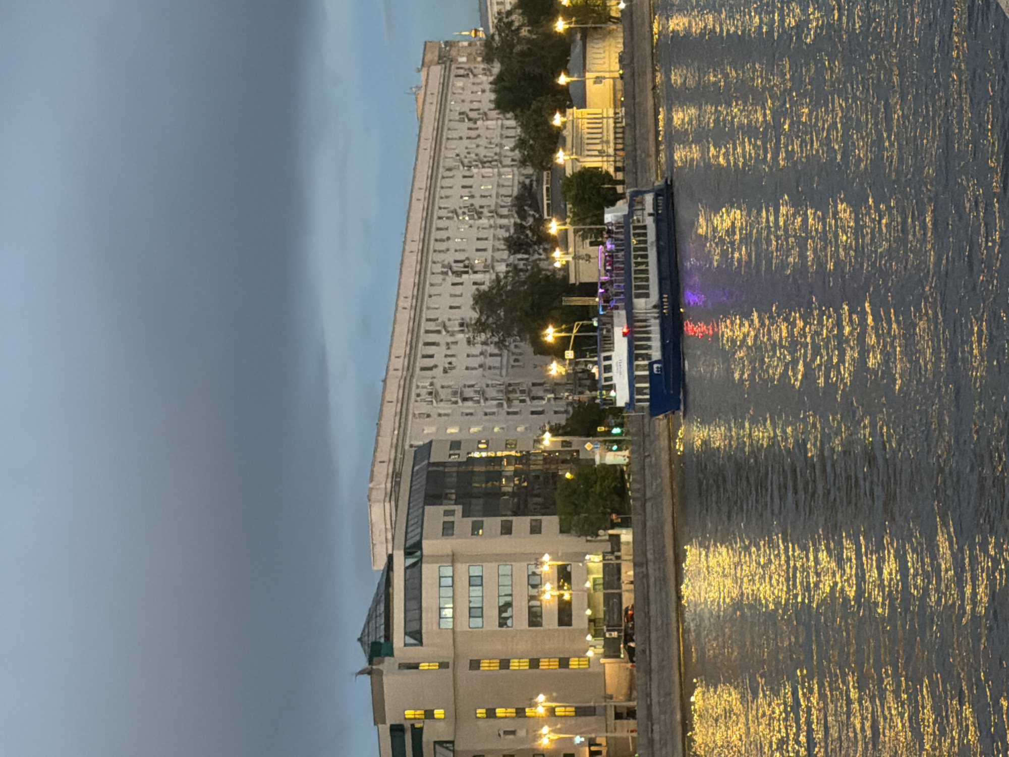 The image captures a view of the Moskva River in Moscow, Russia, at dusk. A white and blue passenger boat, illuminated with colored lights, cruises on the water. Reflections of streetlights and building lights shimmer on the river's surface. Along the embankment, a multi-story building with many windows and lights is visible, adjacent to a longer, pale-colored building with a distinct architectural style. Trees are interspersed between the buildings and along the riverbank. The sky is overcast, with a gradient of gray and blue tones.