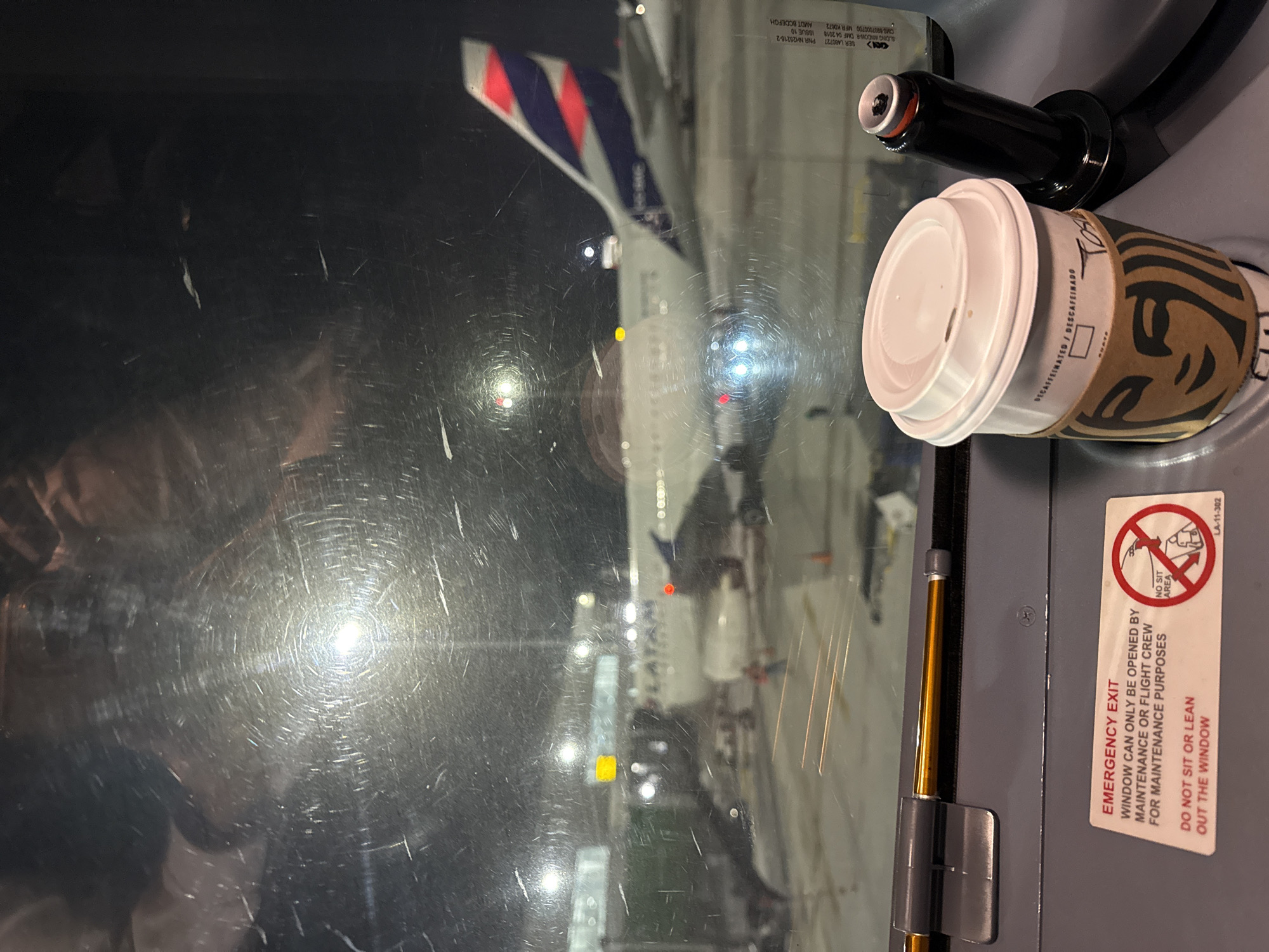 An exterior view from an airplane cabin at night shows a LATAM Airlines Boeing 767 aircraft parked at a gate at Jorge Chávez International Airport in Callao, Peru. The aircraft's tail, emblazoned with the LATAM logo, is visible. The airport tarmac is illuminated by ground lights, and a jet bridge extends towards the plane. In the foreground, on the interior side of the cabin window, a Starbucks coffee cup sits next to a black object, possibly a speaker or microphone. A red and white "EMERGENCY EXIT" sticker with a "NO SIT AREA" symbol is affixed to the interior paneling. The window itself is heavily scratched, creating a diffuse and distorted view of the exterior scene.