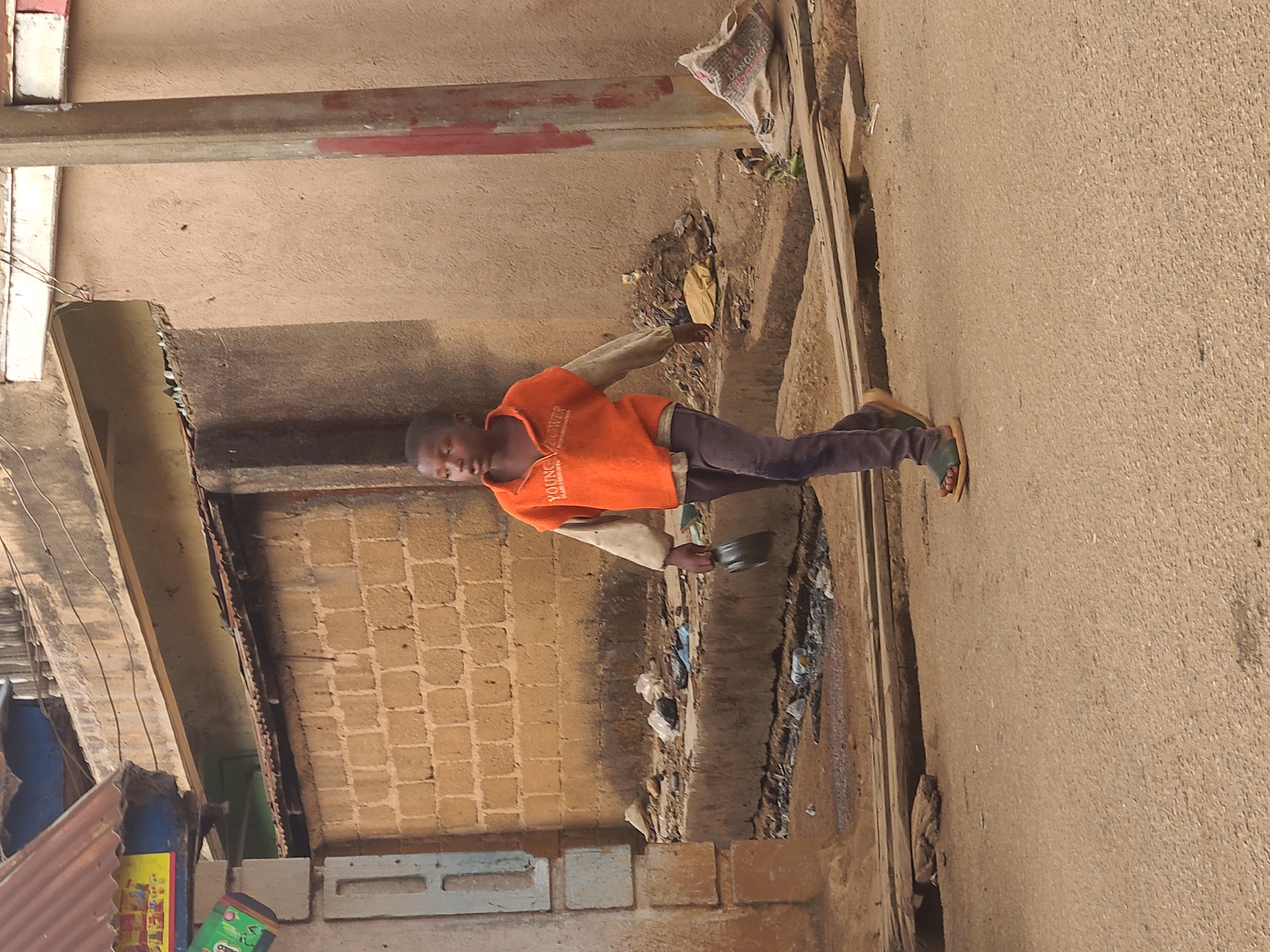 A young boy stands on a street in Jos, Nigeria. He wears an orange shirt, dark pants, and sandals. He is holding a small, dark-colored bowl. To his left is a brick wall and a small structure. To his right is the street. There is debris and litter in the gutter along the side of the street.