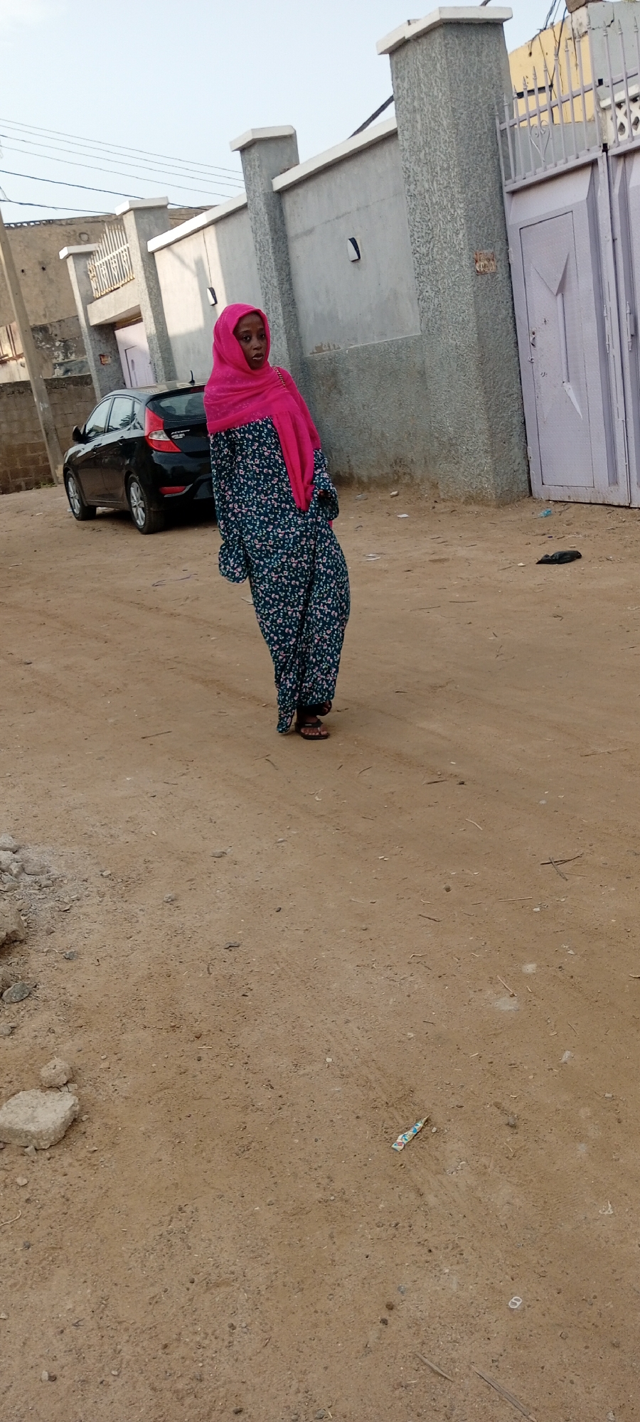 A young woman wearing a pink hijab and a dark floral print dress stands on a dirt road in the city of Kano, Nigeria. She is facing slightly to her left and looking towards the viewer. Behind her, a black sedan is parked. The background shows a gray concrete wall with decorative pillars and a large light purple metal gate. The ground is covered in sand and small rocks. The sky is overcast.