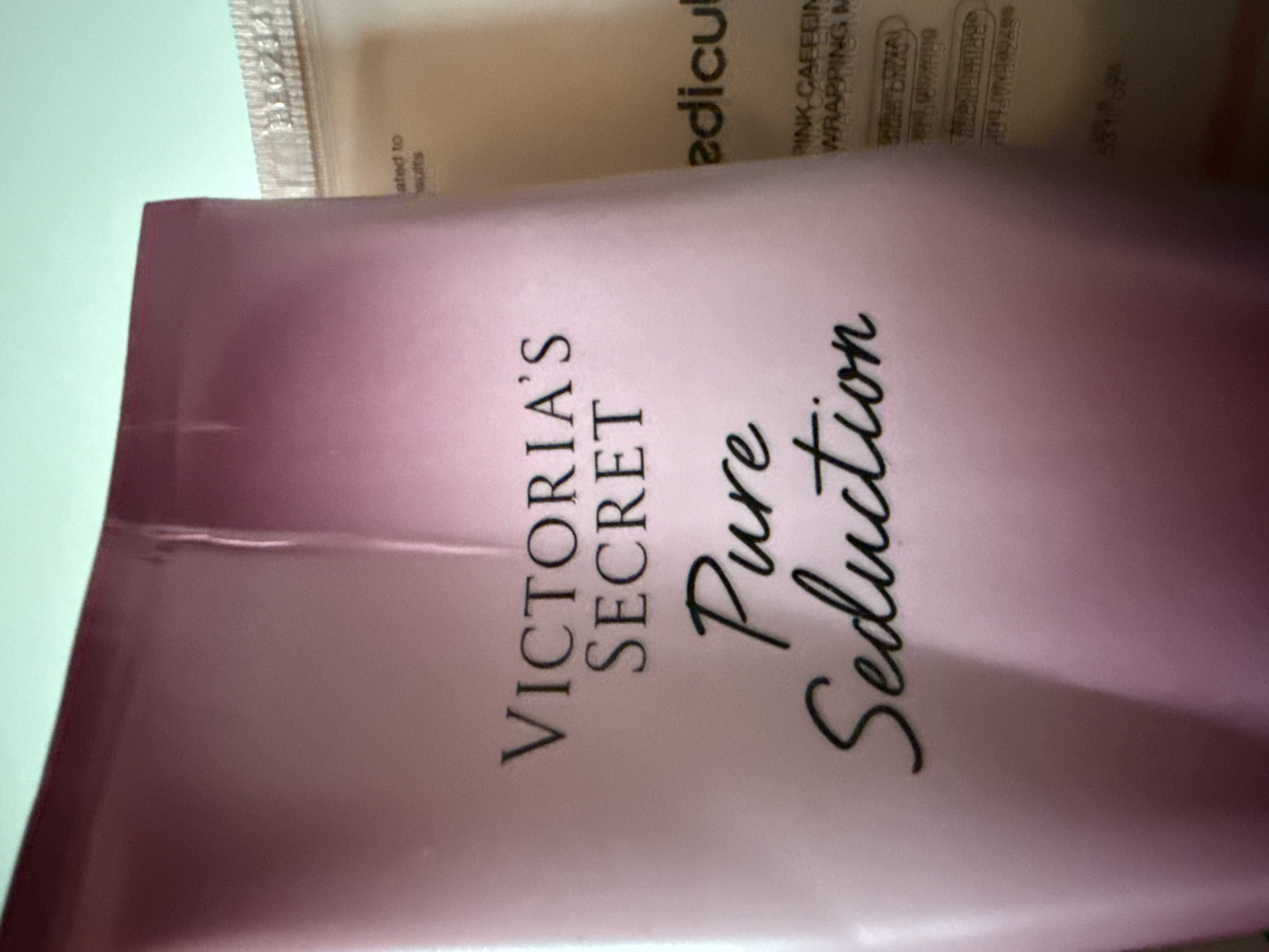 A close-up, eye-level shot shows two bottles of body lotion. The bottle on the left, which is in focus, is pink with a gradient to a darker pink at the top. It has the brand "VICTORIA'S SECRET" in black sans-serif font, and below that, "Pure Seduction" in black script font. The bottle on the right, out of focus, appears to be beige and has black text that is difficult to read. Part of a silver lid or band with a raised pattern is visible on the top right of the image. The background is a light teal color. There are no people or animals in the image, and the setting is not identifiable as Okhtyrka, Ukraine.