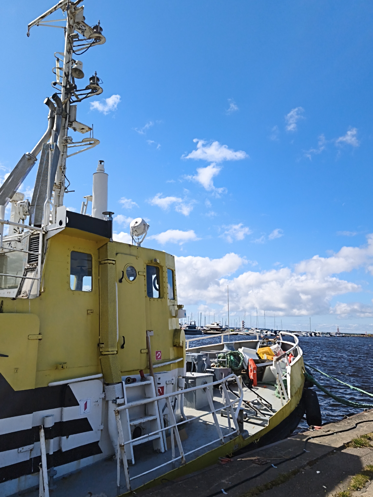 The image shows a yellow and white boat docked in a harbor under a blue sky with scattered white clouds. The boat is the primary subject, with its mast, cabin, and deck visible. The stern of the boat is angled towards the right, with green ropes extending from it to the dock. In the background, other boats and a lighthouse can be seen in the distance. The environment appears to be a port or marina. The presence of a lighthouse and the style of boats suggest a Baltic Sea location. Given the context provided, the location is a harbor in Tallinn, Estonia. There are no people visible in the image.