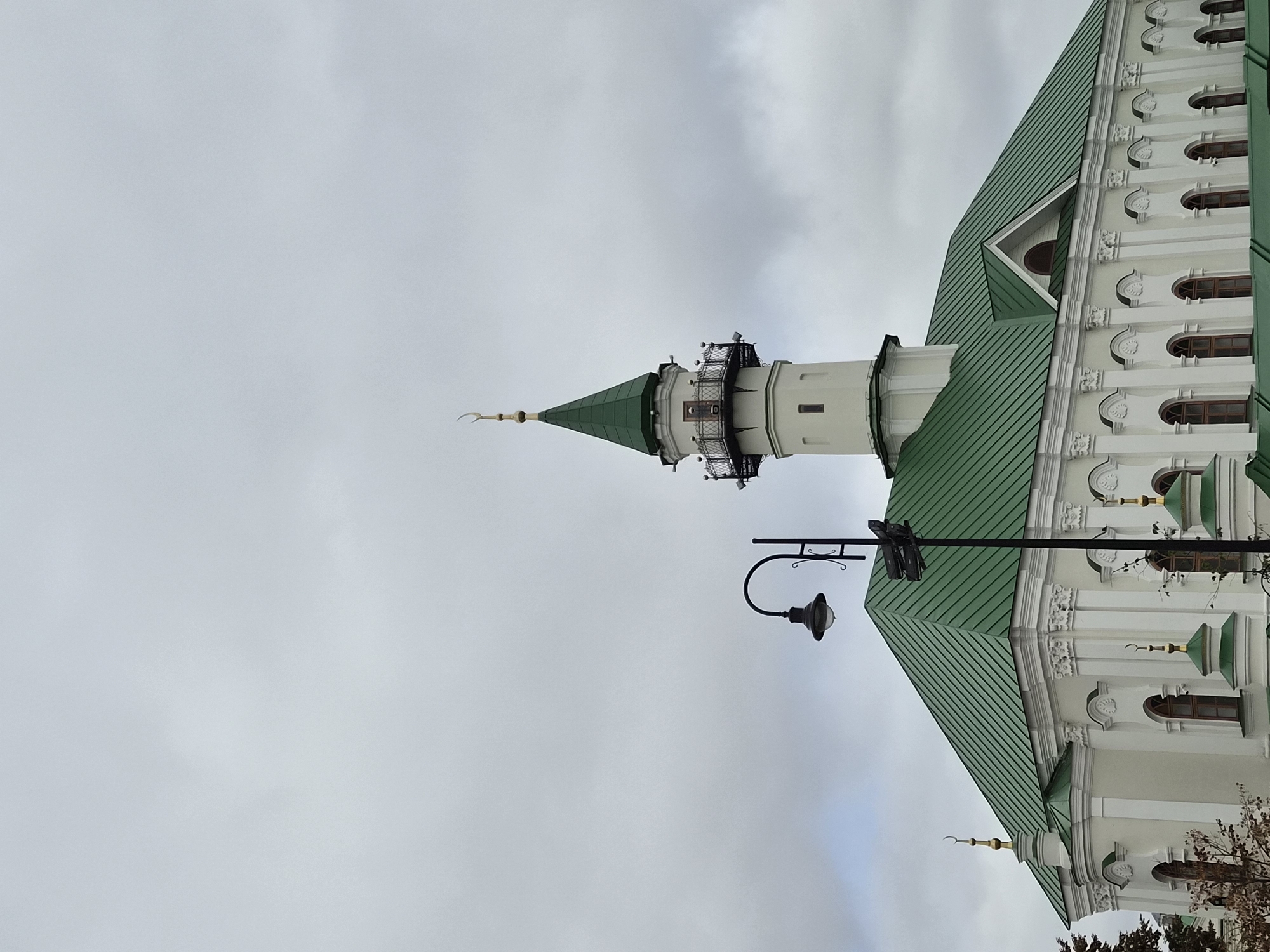 A wide shot captures the Kul Sharif Mosque in Kazan, Russia, under a cloudy sky. The mosque features a prominent white minaret topped with a green spire and a crescent moon, and a sprawling green roof with multiple gables. Decorative white architectural elements adorn the building's facade, which is lined with arched windows. A black lamppost stands in the foreground, to the left of the minaret, with a portion of the mosque's roof visible behind it. The overall scene is static and depicts the exterior of the mosque.