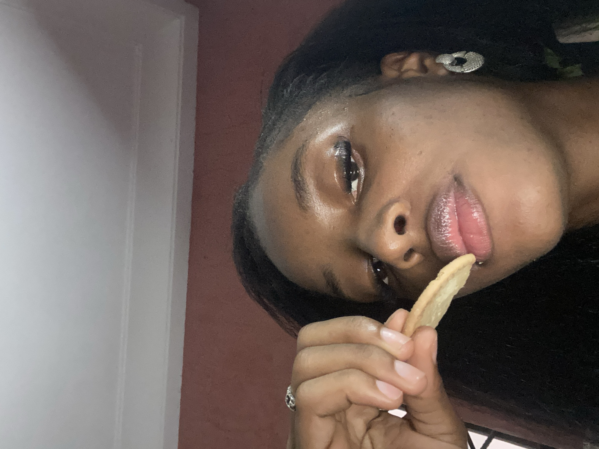 In this close-up, waist-up shot, a young Black woman is captured in a dimly lit indoor setting. Her head is tilted slightly, and she is in the process of eating a single french fry, holding it between her index finger and thumb. Her lips, coated with a shimmering lip gloss, are parted as she brings the fry to her mouth. The lighting highlights the dewy sheen on her forehead and cheekbones, and her eyelashes appear to be enhanced with false lashes. She is wearing a large, silver, hoop-like earring with an intricate, textured design in her right ear. A ring adorns her left hand, visible near her thumb. The background consists of a textured, reddish-brown wall meeting a white ceiling with a subtle molding detail. The overall impression is a casual, personal moment captured in an intimate setting. The context of Jos, Nigeria, cannot be determined from the visual information presented in the image.