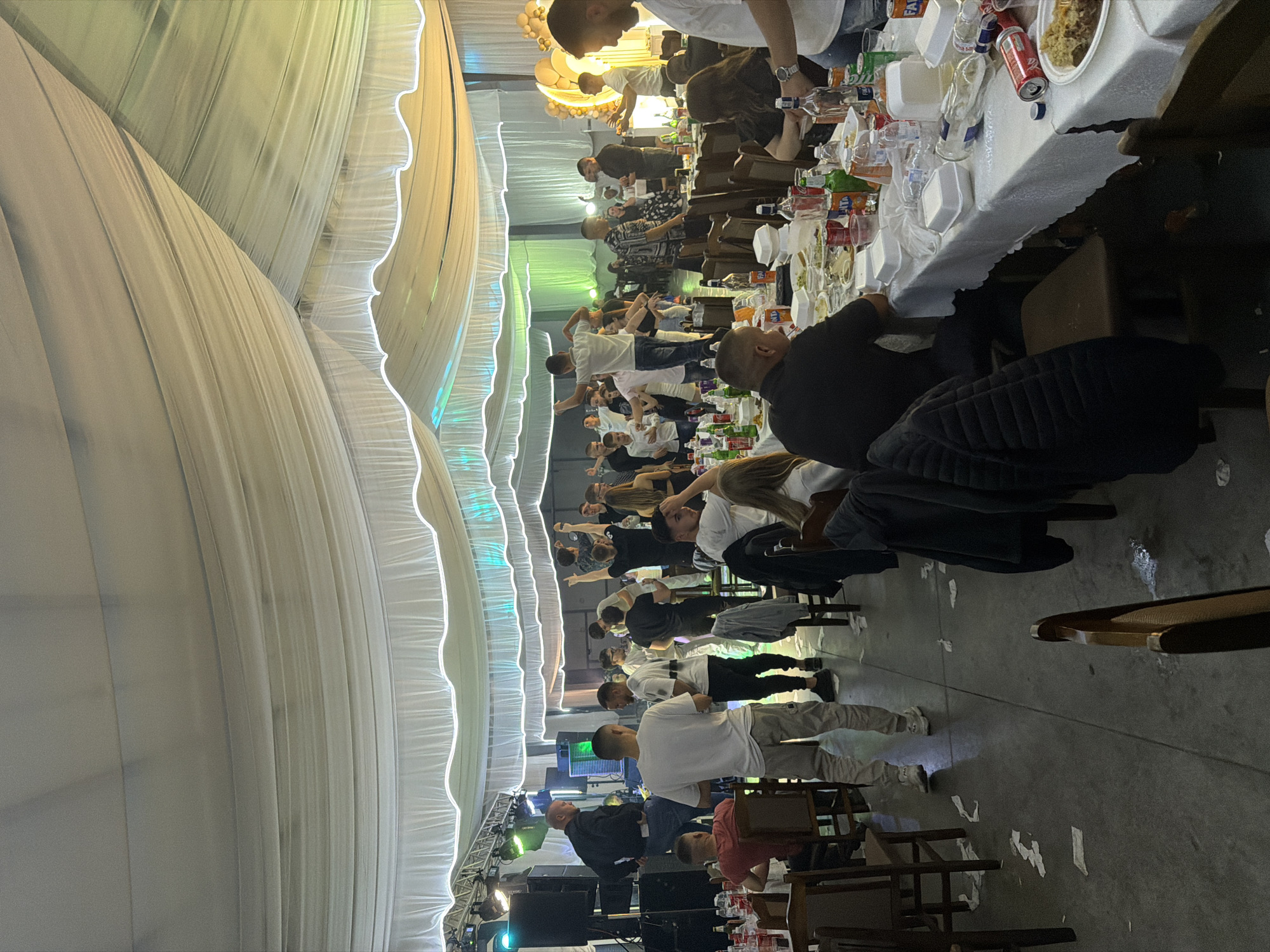 The image depicts a large gathering of people in a spacious tent-like structure, possibly a wedding reception or similar celebration, in Debren, Bulgaria. A long table laden with food, drinks, and disposable tableware stretches across the scene. Individuals are seated and standing, many with their arms raised, possibly in a celebratory gesture. The environment is defined by the white fabric draping of the tent, illuminated by strings of lights. There is stage lighting and sound equipment at the far end of the tent. Scattered tissues and other litter are visible on the floor.