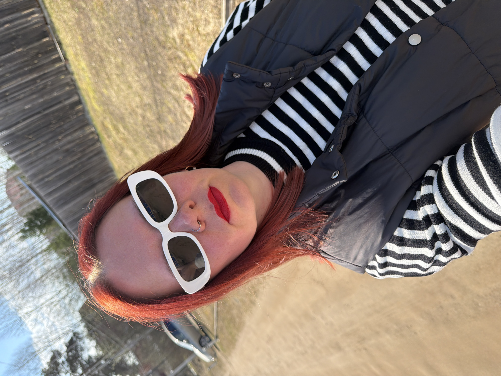 An outdoor portrait captured from a low angle depicts a person with red hair, lying on their side, facing the viewer with their head tilted. The individual is wearing distinctive white, rectangular sunglasses that obscure their eyes, and bright red lipstick. A silver nose ring is visible on their right nostril. Their hair is a vibrant reddish-orange, catching sunlight near the hairline.
The person is dressed in a black and white striped long-sleeved top or sweater, layered with a dark grey or black padded vest that features silver snap buttons. The striped pattern of the top is prominent, offering a stark contrast.
The background is a blurred outdoor scene, indicating a shallow depth of field focusing on the subject. To the left, a wooden fence or structure is partially visible, rendered in muted greys and browns. The rest of the background appears to be a sunlit, grassy or dry field with warm, earthy tones, suggesting a natural and open environment. A faint reflection of trees or sky can be seen in the lenses of the sunglasses.
The lighting suggests a sunny day, with clear skies, providing bright illumination on the subject. There is no visible text in the image. The overall impression is a casual outdoor photo shoot.