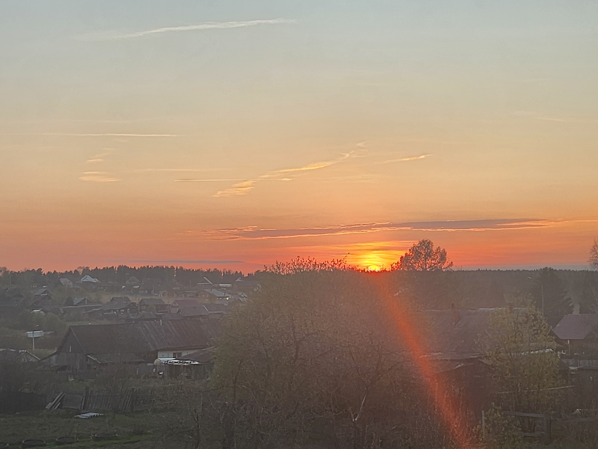 The image shows a sunset over the village of Krasnogorskoye, Russia. The sky is a gradient of colors, from light orange near the horizon to pale blue at the top of the frame. Several contrails are visible in the upper portion of the sky. The sun is setting behind a line of trees, with the rooftops of houses visible in the foreground. Trees and bushes are prominent in the foreground, obscuring some of the houses. A lens flare is visible emanating from the setting sun.