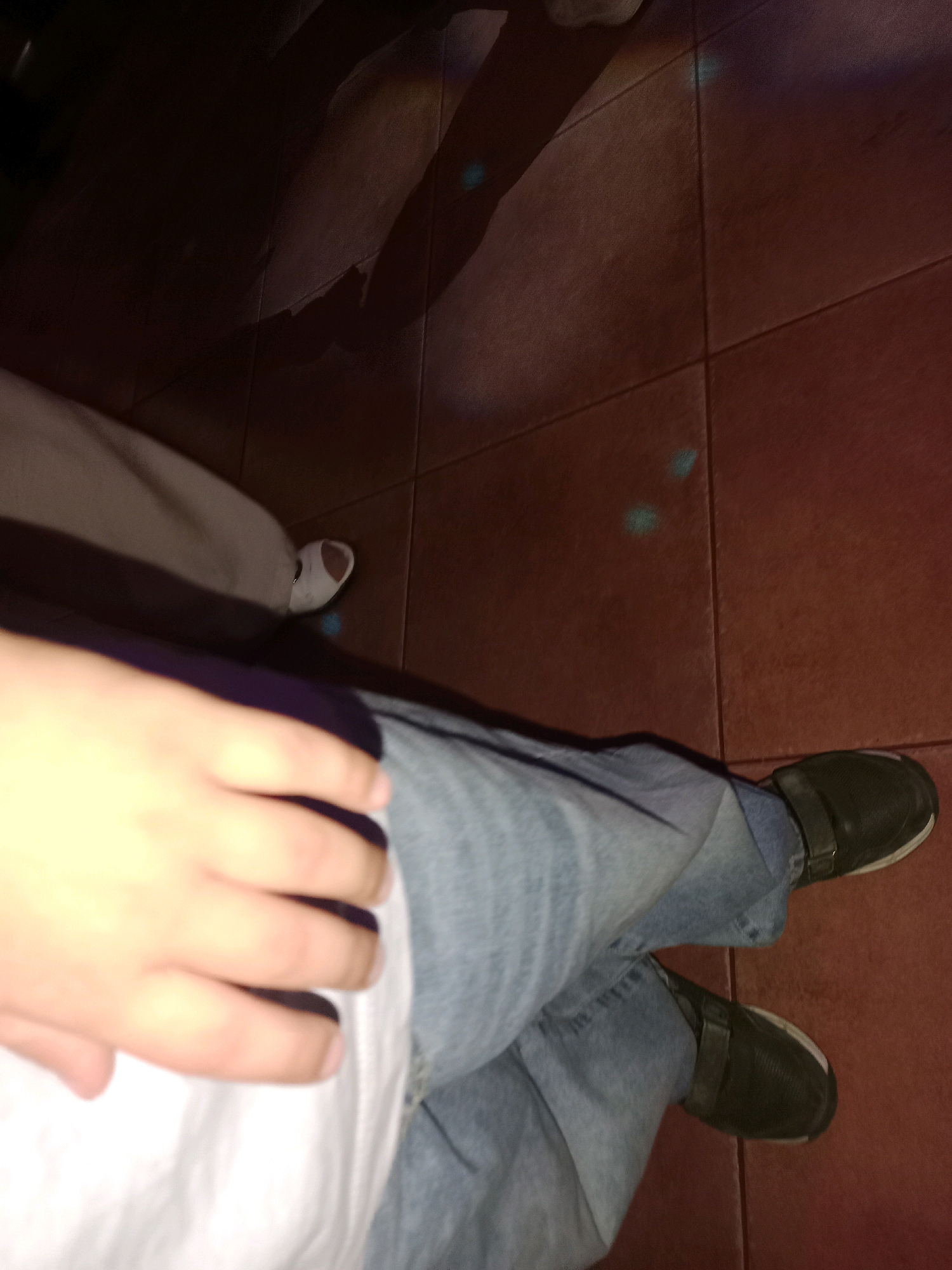 The image captures a low-angle view of a person's legs and feet, standing on a tiled floor. The person is wearing light blue denim jeans and dark green slip-on shoes with a strap across the instep. Their right hand, with visible fingers and fingernails, rests on their left thigh, which is covered by a white shirt or garment. A shadow of another person or object is cast on the floor behind them, suggesting a dimly lit environment, possibly with colored lights reflected on the floor, indicated by faint blue circular patches. The floor appears to be made of large, reddish-brown square tiles. The scene is set within an indoor location in Хацежынскі сельскі Савет, Belarus, though the specific context is not discernible from the visual information provided.