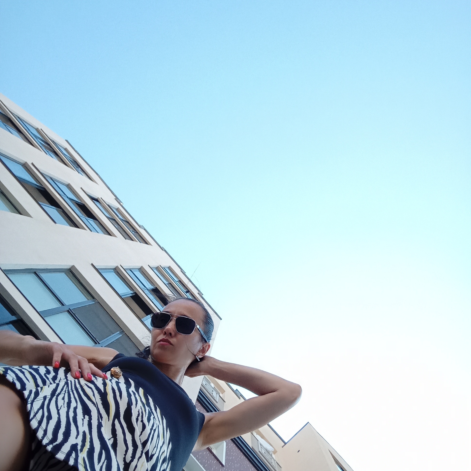 A low-angle shot features a woman wearing sunglasses and a black top with a zebra-print skirt, posed in front of a modern building under a clear blue sky. Her left hand rests on her hip, and her right arm is bent, with her hand behind her neck. The building, characterized by its cream-colored facade and numerous rectangular windows, occupies the left side of the frame. The woman's face is partially visible, looking upwards and to the right. This scene is situated in the city of Pervomaysky, Russia.