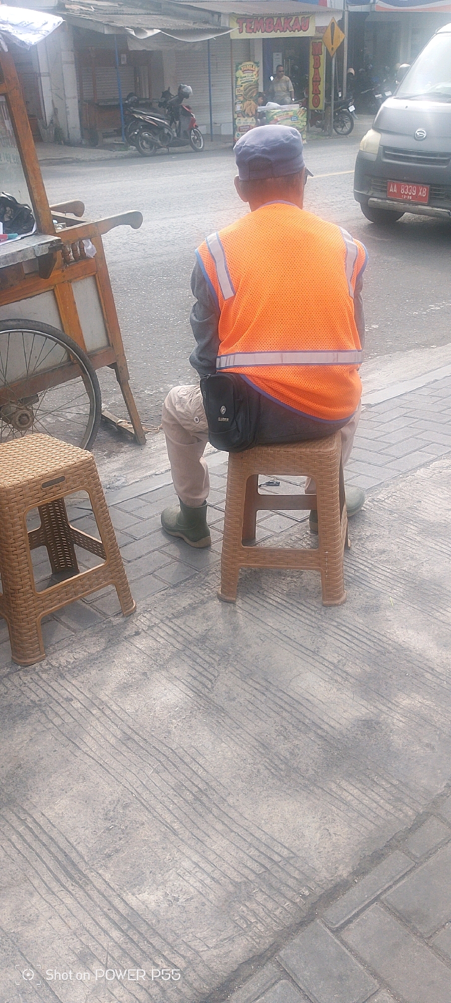 A person wearing a blue cap and an orange reflective vest sits on a brown plastic stool, facing away from the camera. The person is wearing tan pants and dark green boots. A black pouch is attached to the person's waist. To the left of the person is a brown wicker stool, and to the right is a brown plastic stool. In the background, a street is visible with several motorcycles and a dark-colored car. There are also street vendors and shops. The ground is paved with gray interlocking blocks and concrete.