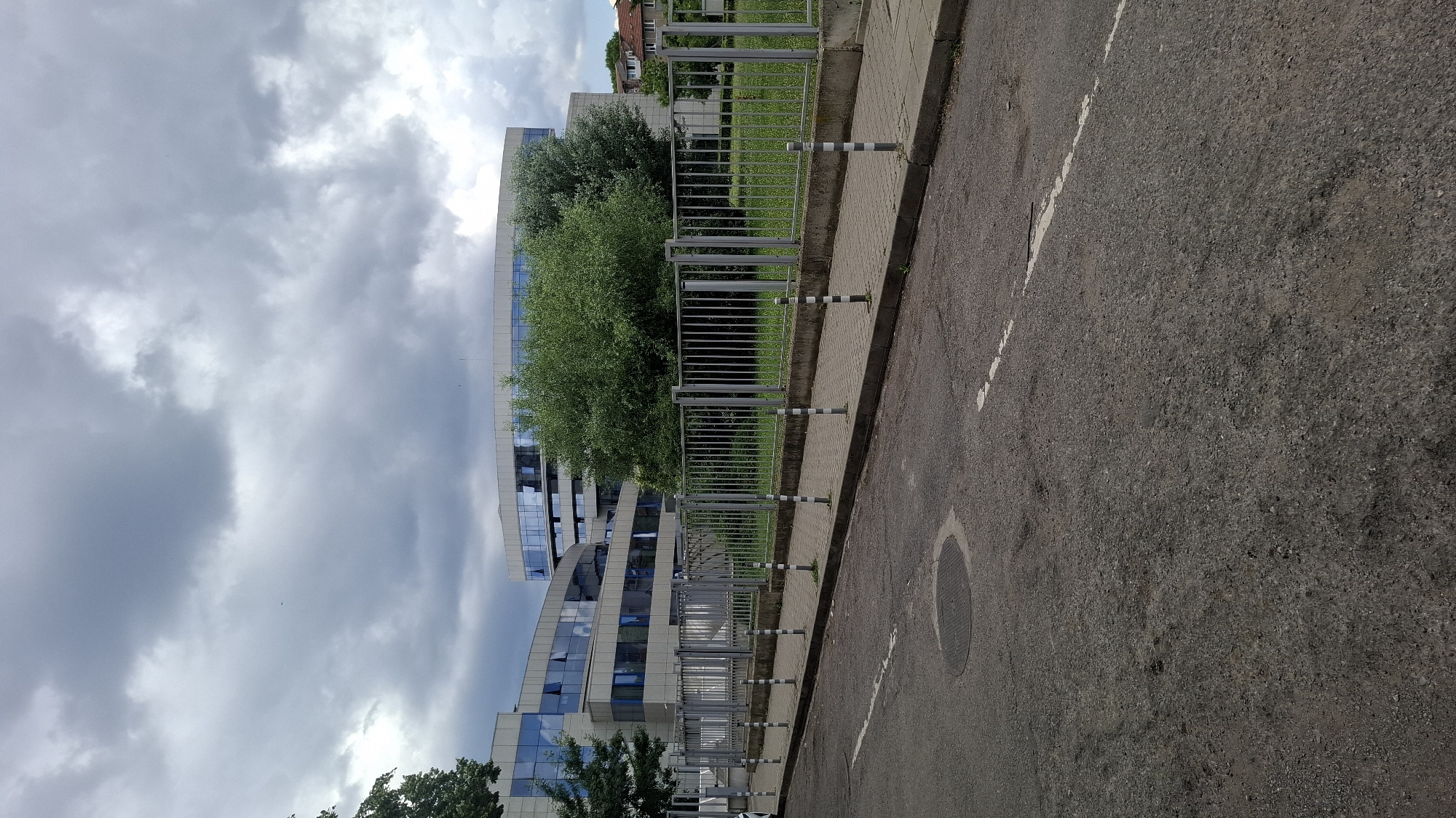 The image depicts a street-level view of a modern, multi-story building with a curved facade and large blue-tinted windows, likely an office building. A metal fence separates the sidewalk from a grassy area in front of the building. The sky is overcast with gray clouds. The street in the foreground is asphalt with some wear and tear, including a manhole cover. The scene appears to be located in Sofia, Bulgaria, based on similar architectural styles and urban settings found in the city. No people or vehicles are visible in the image.