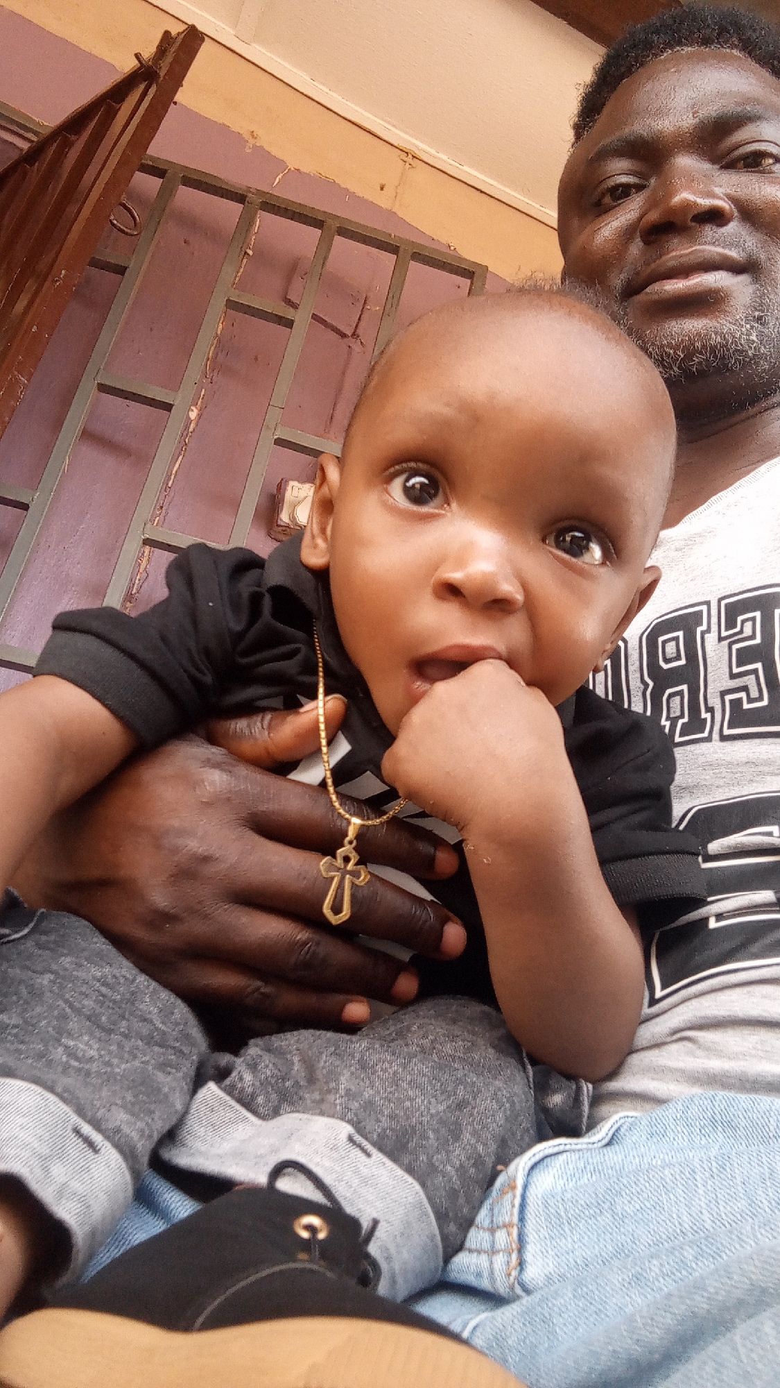 A photograph shows an infant being held by an adult. The infant is light brown skin wearing a black shirt and a gold cross necklace. The adult is dark brown skinned, with gray-flecked facial hair, and a gray and white patterned t-shirt. The background features a purple wall with a rusted metal security bar. The photograph was taken in Sabon Tasha, Nigeria.