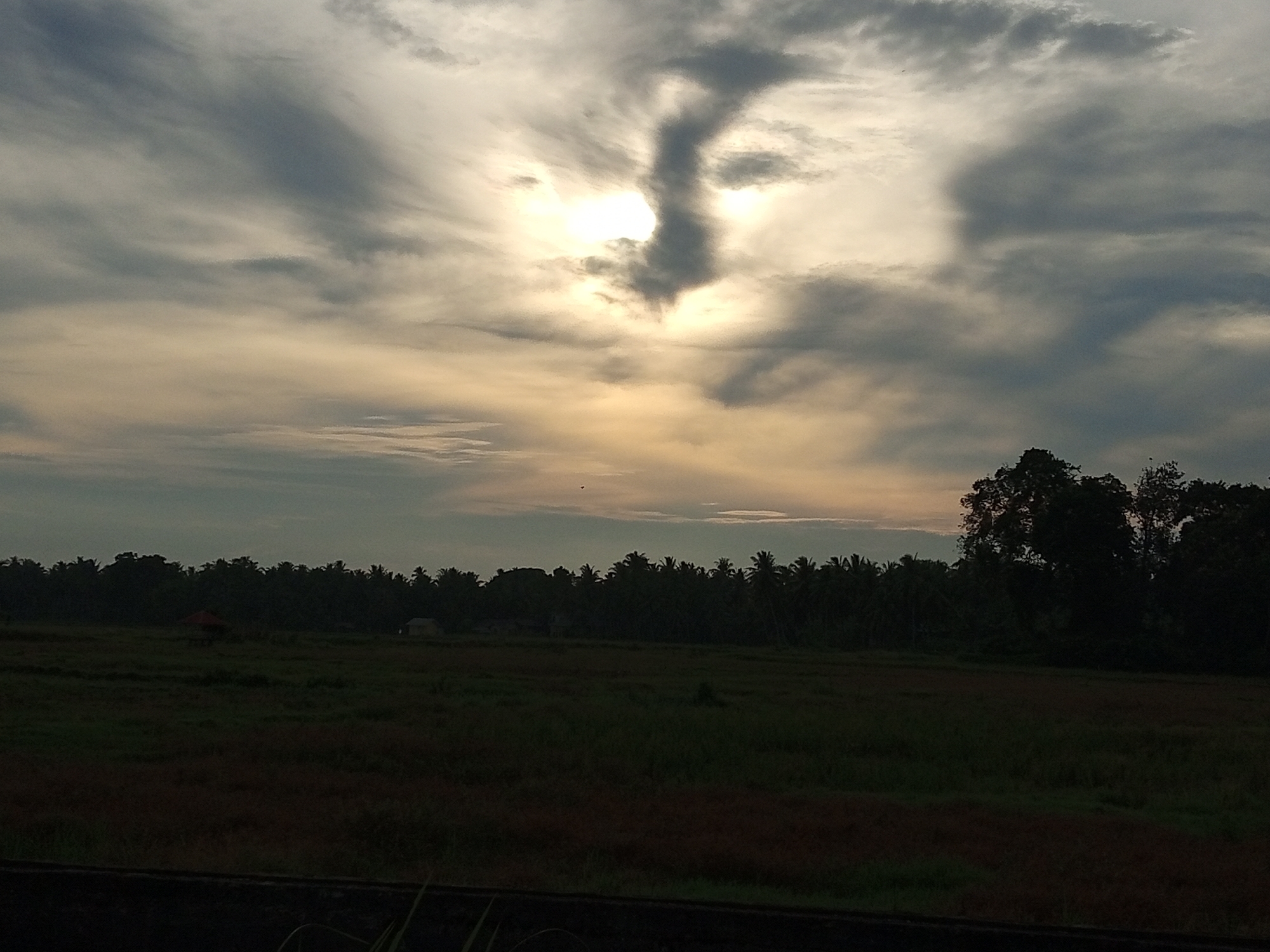 The image depicts a wide shot of a rural landscape in Cotjrat, Indonesia, during what appears to be either sunrise or sunset. The sky is filled with dramatic clouds, with the sun peeking through, casting a soft, warm glow. A prominent, dark cloud formation in the center of the sky resembles a human figure, adding a striking element to the scene.
Below the sky, a dense line of dark green trees forms the horizon, suggesting a forested area. In the foreground, a vast, open field stretches out, appearing to be a cultivated area, possibly rice paddies, given the dark, reddish-brown and green patches of vegetation. A small, red-roofed structure, possibly a hut or gazebo, is visible within the field on the left side of the image, along with a smaller, light-colored building further back. The overall impression is one of tranquility and natural beauty in a rural Indonesian setting.