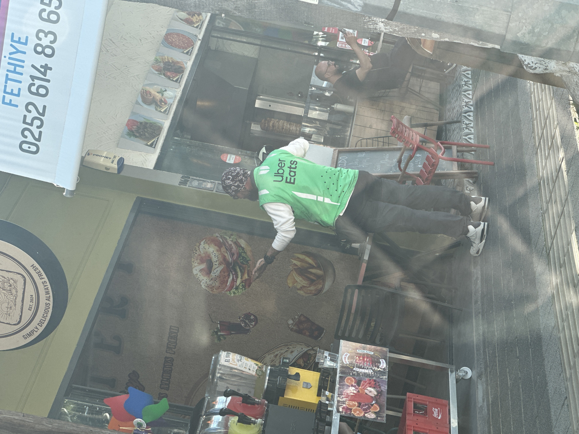 This street-level photograph, taken in Fethiye, Turkey, captures an Uber Eats delivery driver outside a food establishment during what appears to be a bright, sunny day. The main subject is a man wearing a distinctive bright green Uber Eats vest over a white long-sleeved shirt, with a patterned head covering and dark trousers. He stands in front of the shop window, looking down, possibly checking his phone or a delivery device, suggesting he is either awaiting or collecting an order.
Inside the shop, another man, possibly a chef or employee, is visible through the window, behind a glass counter that features a large döner kebab skewer. He wears a dark t-shirt and glasses, seemingly engaged in food preparation or serving.
The shop exterior displays various food decals on its window, including a large bagel burger, a bowl of fries, and drink illustrations. Below the window, several drink dispensers and red Coca-Cola crates are visible. Stacked red plastic chairs sit near a small chalkboard menu outside.
Visible text provides location and business details: A white banner at the top left advertises "FETHİYE 0252 614 83 65," indicating a local phone number. A round sign on the shop's facade reads "SIMPLY DELICIOUS ALWAYS FRESH EST. 2024." Inside, small framed images on the wall display food items like "Iskender," "Ezogelin," "Dürüm," and "Adana Kebap." The chalkboard menu lists items such as "Mercimek," "Tavuk Suyu," "Ayran," "Şalgam," "Su," and "Sodas," with prices in Turkish Lira. A small security system device with "PRONET 444 1911" is also mounted near the banner. The vibrant green of the Uber Eats vest contrasts with the muted tones of the street and shop facade, highlighting the modern delivery service in a traditional urban setting.