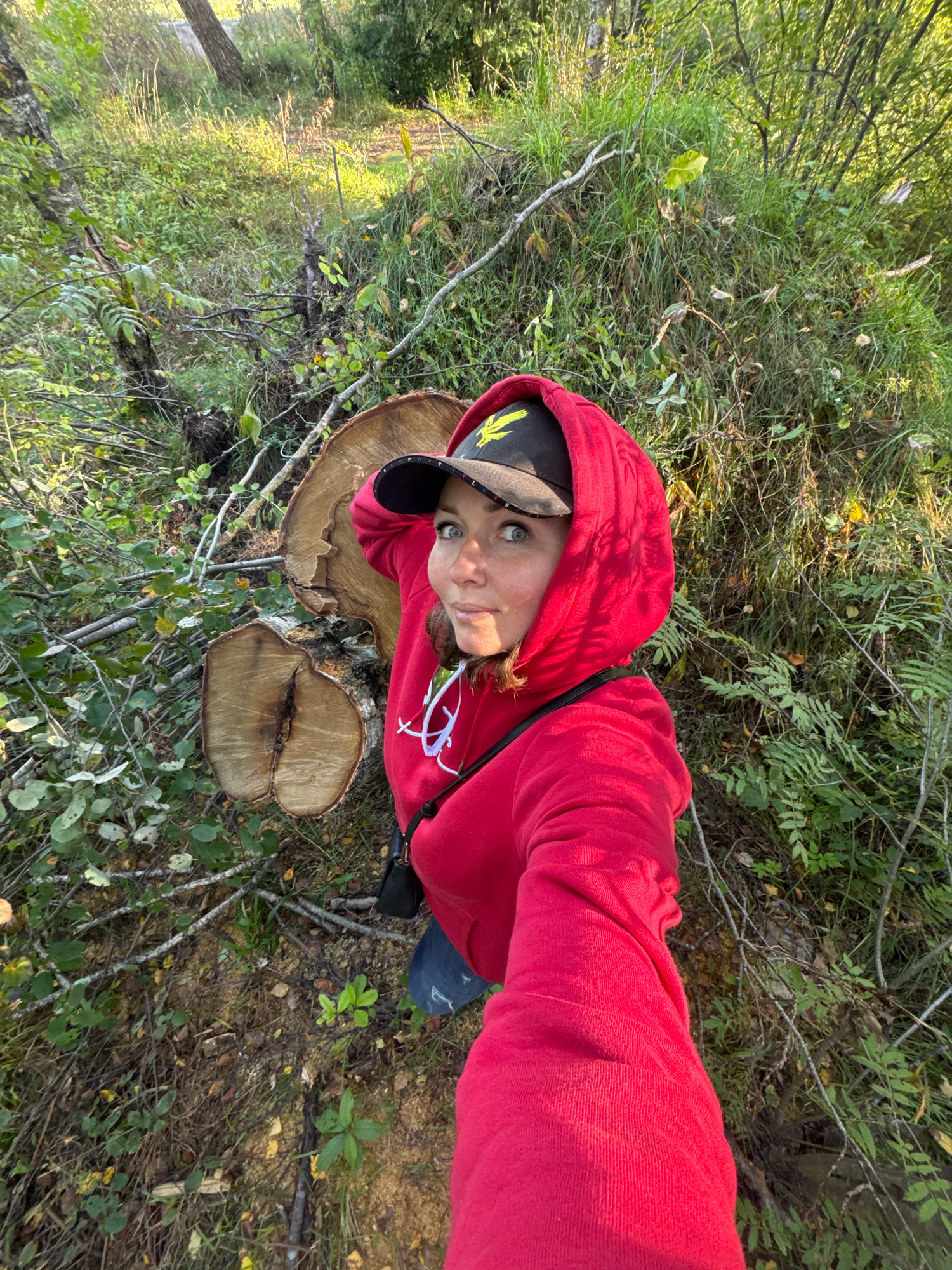 Innolovo, Russia. A woman in a red hooded sweatshirt and a black baseball cap is taking a selfie in a wooded area. She is standing next to a large, freshly cut tree trunk with visible rings. The area is overgrown with green vegetation and branches. The sunlight is casting shadows on the ground, indicating it is likely late afternoon or early morning. The woman has her left hand on the brim of her cap, and her gaze is directed at the camera. The scene appears natural and untamed.