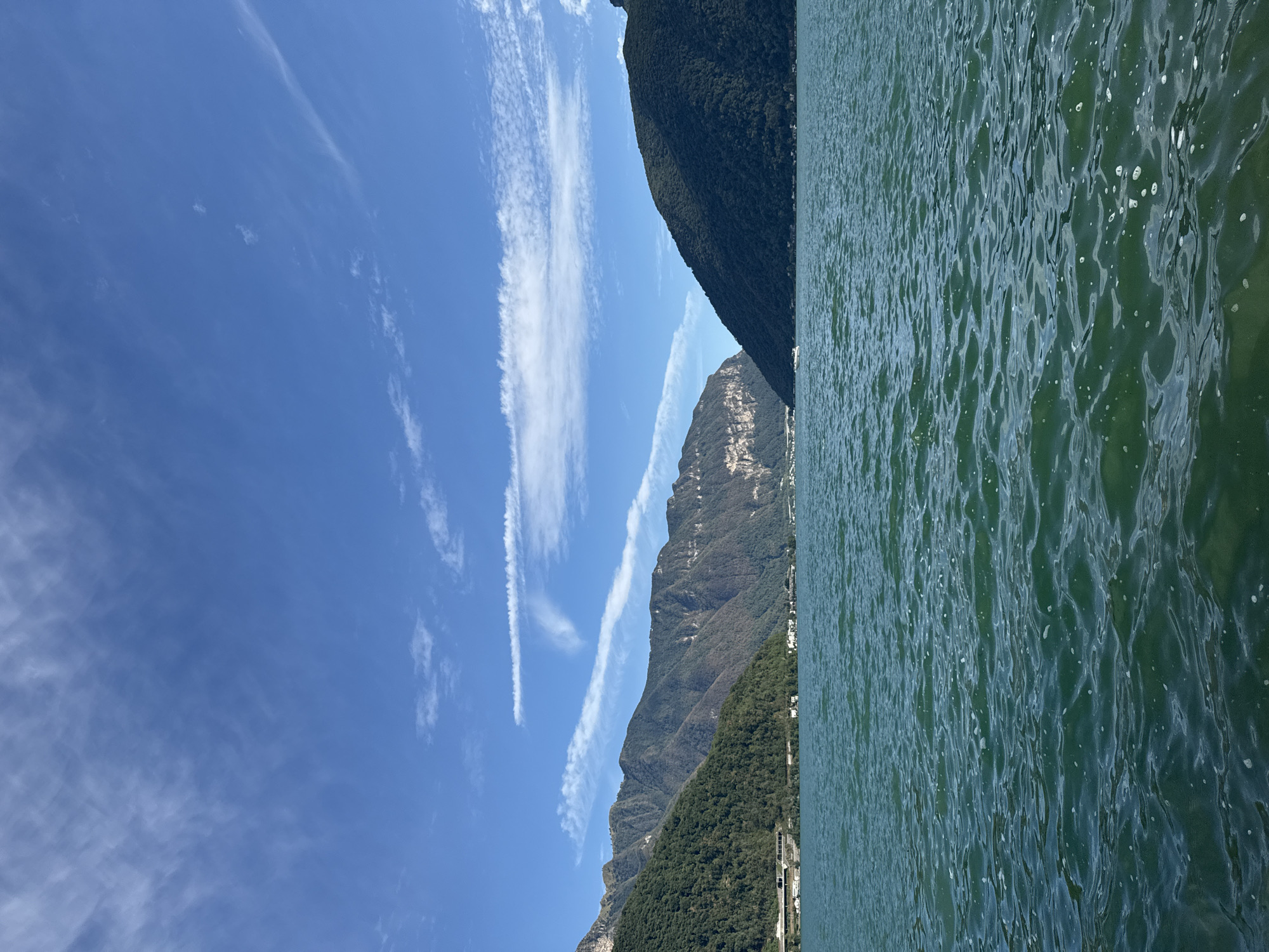 This is a photograph of Lake Lugano, taken from the water, looking towards the town of Melide, Switzerland. The water in the foreground is a deep teal color with visible ripples and some scattered foam. In the mid-ground, the shoreline of Melide is visible, with low-rise buildings nestled at the base of tree-covered mountains. A white boat is visible on the water in the distance, heading towards the right side of the frame. The mountains on either side of the lake are covered in dense green foliage, with rocky outcrops visible on the left mountain. The sky above is a clear blue, with thin, wispy white clouds stretching across it. The overall impression is one of a serene, natural landscape under a bright, sunny sky.