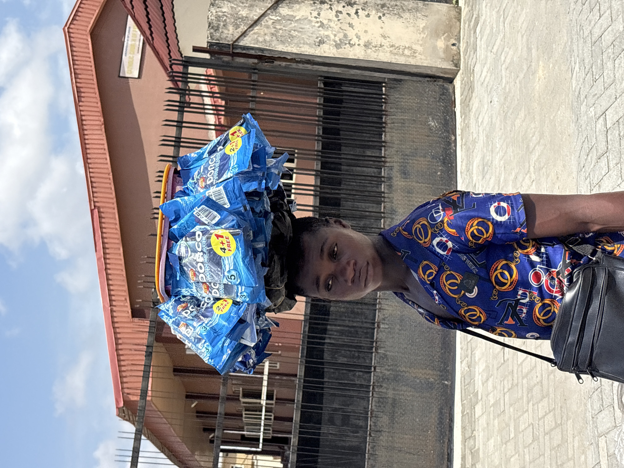 A medium, eye-level shot, rotated ninety degrees, shows a person, possibly a young man, carrying a large stack of "Dorco Twin Blade" razor packages on their head. The razor packages are predominantly blue with yellow and white accents. The person is wearing a blue short-sleeved shirt with a gold and red design, and a black bag is slung across their body. The setting appears to be urban, with a building featuring a red-tiled roof and a barred metal gate visible in the background. A light-colored paved sidewalk is underfoot. The location is Igboefon, Nigeria. The sky is visible in the upper left corner, partially obscured by the building.