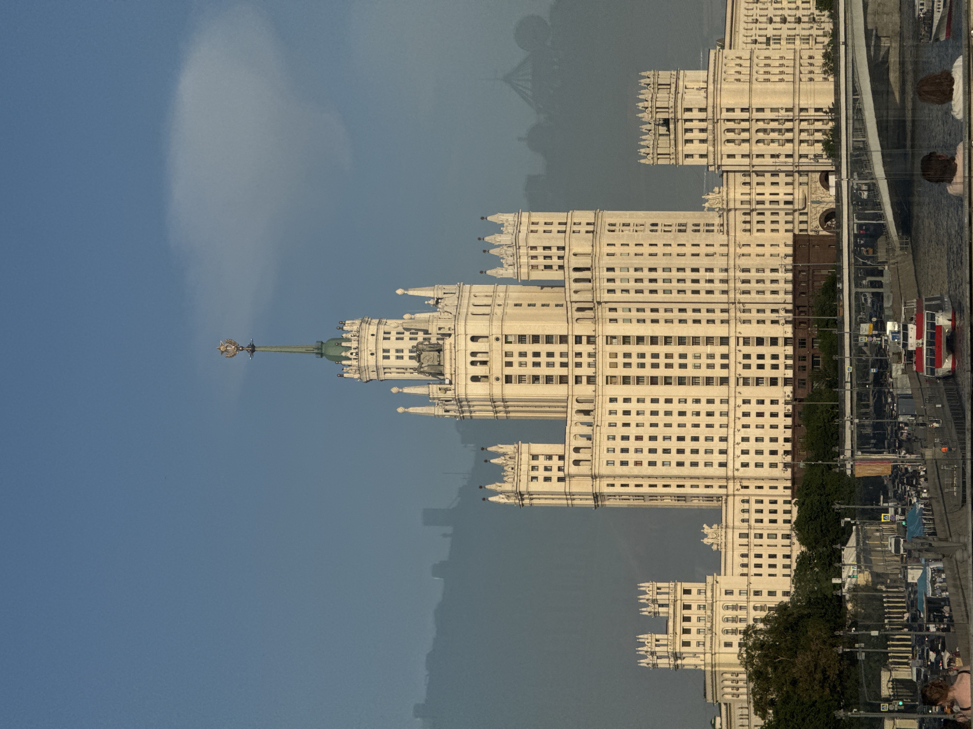 The image depicts the Kotelnicheskaya Embankment Building, one of the Seven Sisters skyscrapers in Moscow, Russia, viewed from an elevated position. The building is tall, multi-storied, and predominantly white or light-colored, featuring Stalinist architecture with ornate detailing. The spire is topped with a star. The sky is clear blue. Some people and vehicles are visible on the right side of the image in what appears to be a reflection. A bridge or elevated structure is visible near the buildings, partially reflecting light.