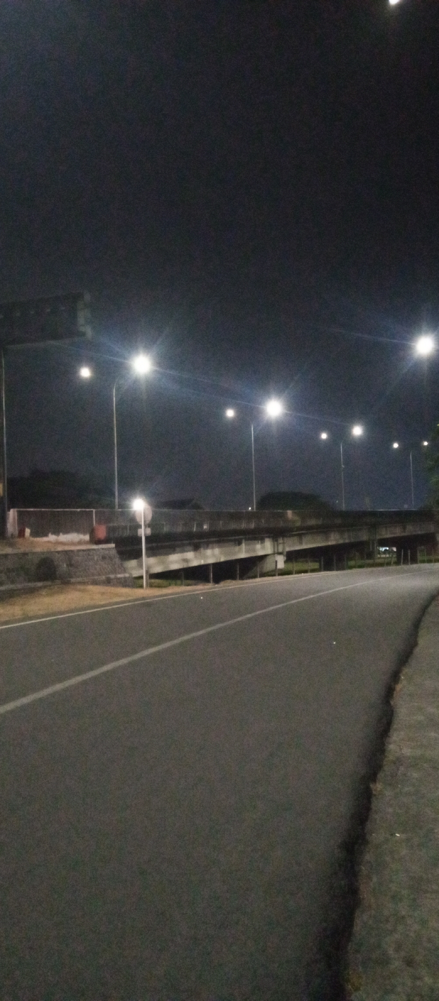 A wide asphalt road curves to the right in the foreground, with a white line dividing lanes. In the background, a concrete overpass spans across the scene, supported by pillars. Streetlights illuminate the overpass and the surrounding area, casting long shadows. The sky is dark, indicating nighttime. The scene appears to be a quiet road in an urban or semi-urban setting, possibly near a highway interchange. The overall impression is one of an empty road at night, lit by artificial lights.