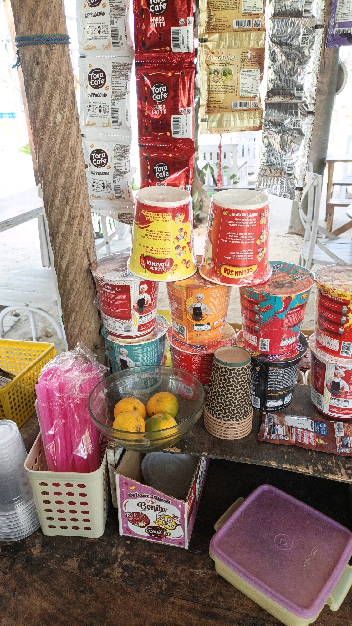 This image captures a street stall display in Cikal, Indonesia, showcasing a variety of instant food and beverage products. The stall is constructed from wooden planks and a thick rope acts as a primary support. Products are displayed on multiple levels, with hanging sachets of "Tora Cafe" cappuccino and chocolate latte, as well as other bagged items with Indonesian text. Below these, stacked plastic cups of instant noodles are prominently featured, with different brands and flavors visible. A clear glass bowl contains a few oranges. Adjacent to the bowls are pink plastic straws in a white plastic basket and a stack of disposable cups. A cardboard box with an image of a woman and text in Indonesian, potentially for a beverage mix, sits on the lower shelf. Other items include small packets of food or snacks and a purple plastic container. The background suggests an outdoor setting, possibly near a beach or recreational area, with glimpses of white furniture and foliage. The overall impression is a bustling, informal market stall designed to attract customers with readily available snacks and drinks.