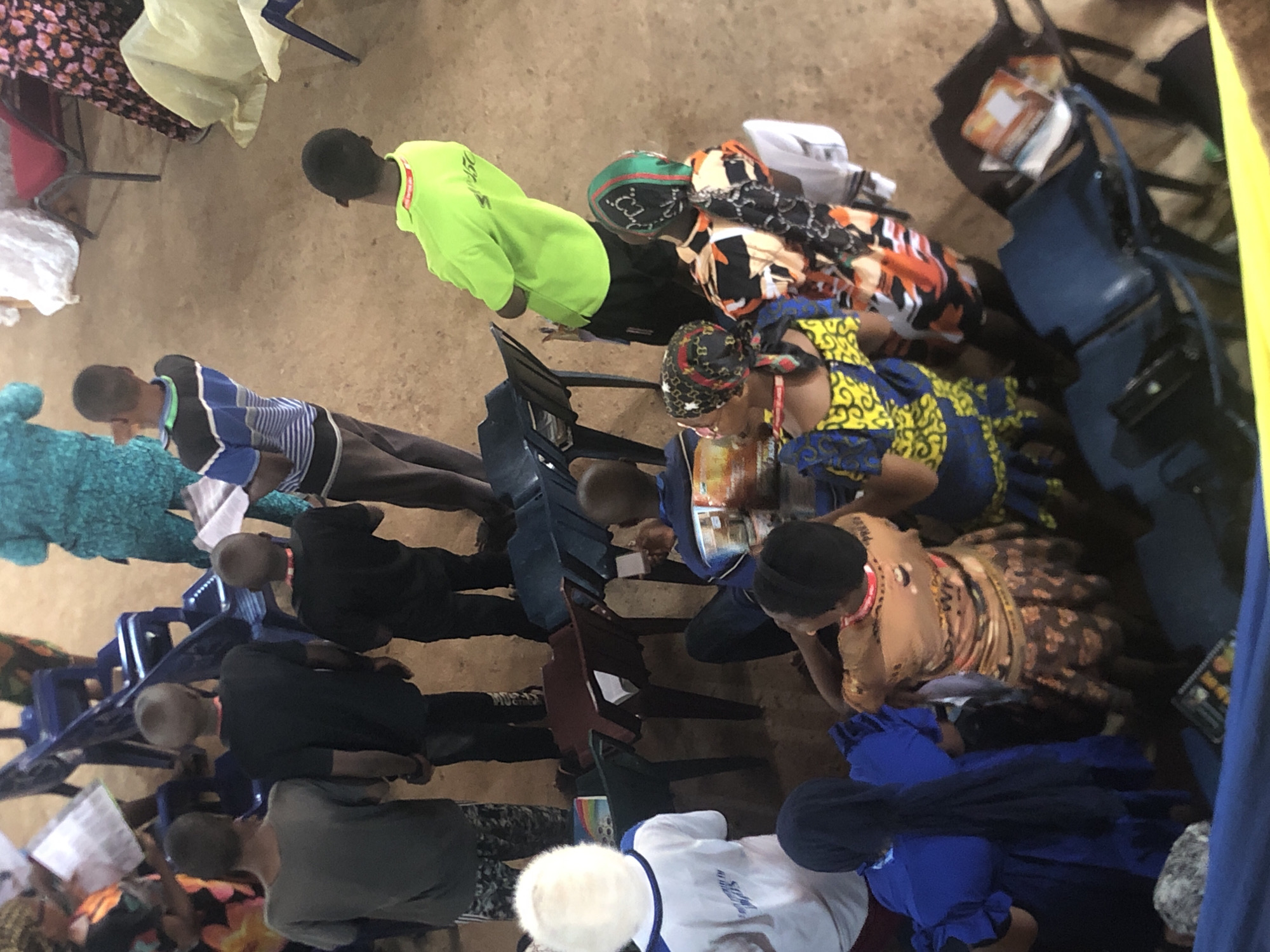 A high-angle shot shows a group of people seated in rows of chairs in what appears to be an indoor space. The setting is in Obosi, Nigeria. The people are likely attending an event or service, perhaps a religious gathering, as some are seen reading from books or holding papers.
The attendees are dressed in a mix of clothing, including traditional African prints and western-style attire. One individual is wearing a bright neon green long-sleeve shirt. Some individuals have head coverings or hats.
The chairs are mostly blue plastic chairs. Several bags, purses, and other personal belongings are placed on the ground. The floor is an unpaved surface, possibly dirt or concrete. The lighting appears to be natural, with daylight coming in from the side.