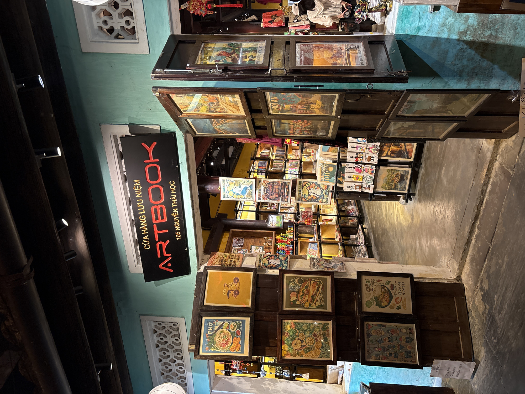 The image shows the entrance of an art book store named "ARTBOOK" located at 105 Nguyễn Thái Học street in Hội An Ward, Vietnam. The store's facade is painted in a light blue color, and there are decorative, white-framed, patterned windows. A black sign with the name of the store is vertically oriented and written in red font. The store's interior displays numerous framed posters and prints arranged on shelves and walls, including framed posters of Vietnamese food. The sidewalk in front of the store is visible, suggesting an outdoor setting, and there is a sign in Vietnamese on the floor at the very edge of the frame.