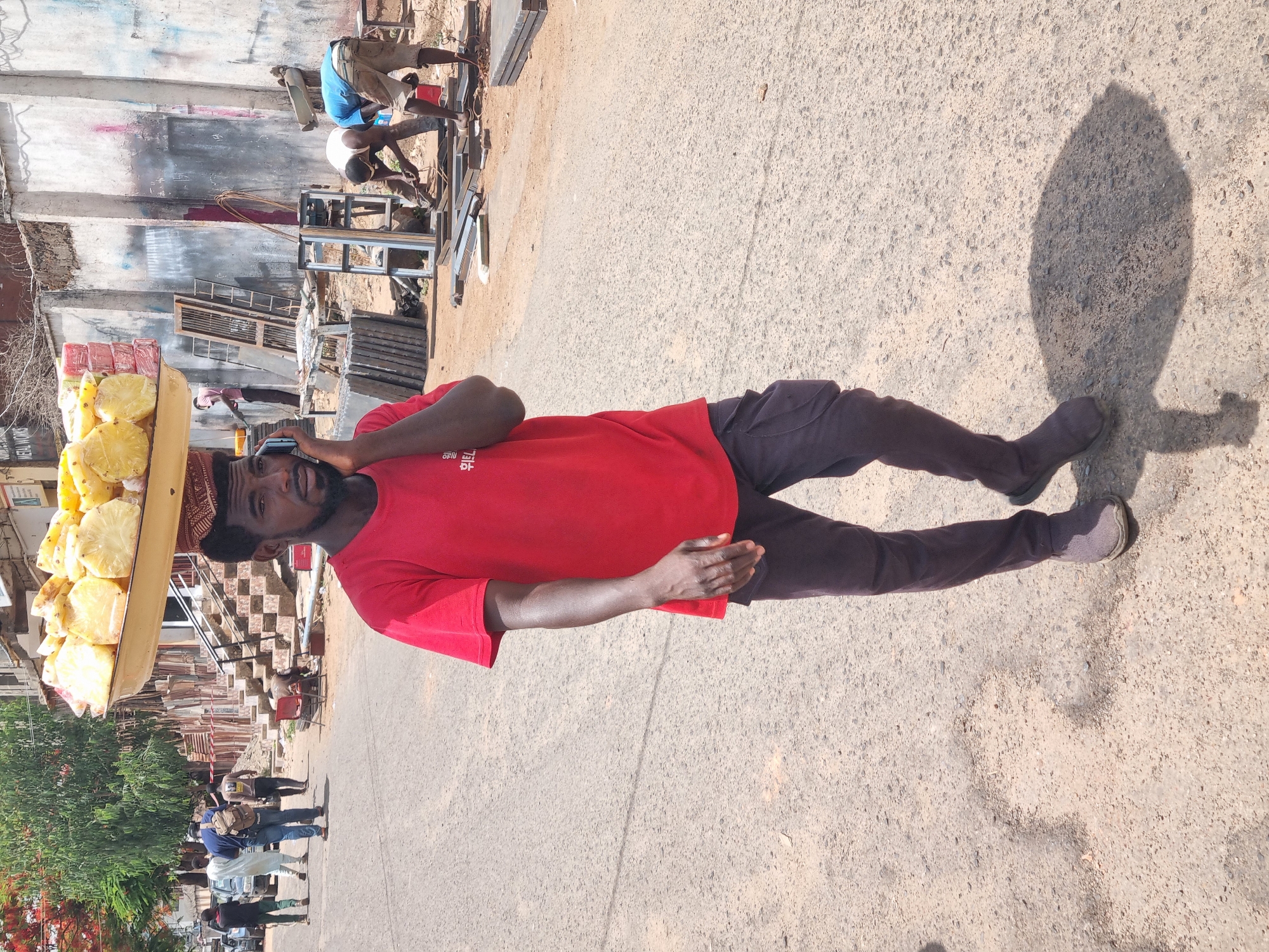 The image shows a man wearing a red shirt and dark trousers, standing on a paved road in Jos, Nigeria. He is balancing a tray of cut pineapple and possibly other fruits on his head while holding a cell phone to his ear. His shadow is cast to his right. In the background, there are buildings, some under construction, and other people walking along the road. Further back, two men are working with metal, possibly welding or cutting. The environment suggests a busy urban street scene, with commercial or residential buildings and industrial activities occurring simultaneously.