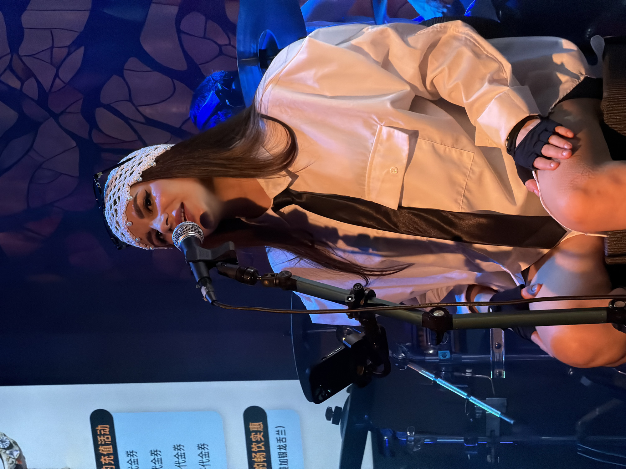A young woman with long dark hair and a white crocheted headband adorned with pearls sings into a microphone. She is wearing a white button-down shirt, a black tie, and fingerless black gloves. The setting appears to be indoors, possibly a performance venue, with a dark blue, abstract patterned backdrop. The lighting is dramatic, with blue hues and spotlights illuminating the singer. In the foreground, there are signs with Chinese text, which appear to be advertisements for "Recharge Activities," "Vouchers," and "Drink Discounts." The woman's expression is focused and engaged, suggesting she is in the midst of a performance. The image is taken from a slightly low angle, emphasizing the singer's presence.