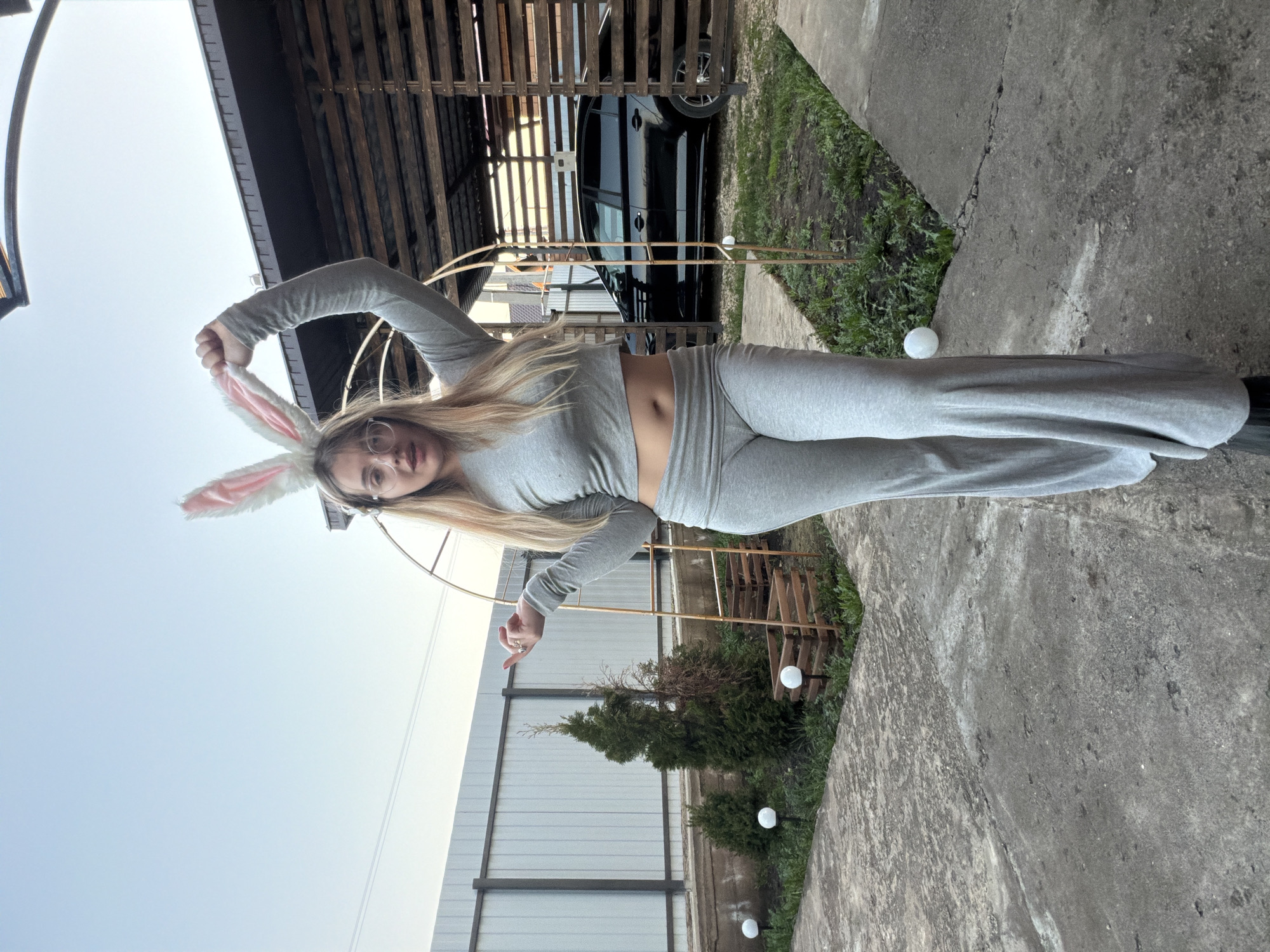 The image, taken in Большая Шильна, Russia, shows a woman wearing a gray long-sleeved crop top and matching pants, and bunny ears. She is posing outdoors near a fence, greenery, and outdoor lighting. Behind her is a structure of wood and a black car. The ground is concrete.