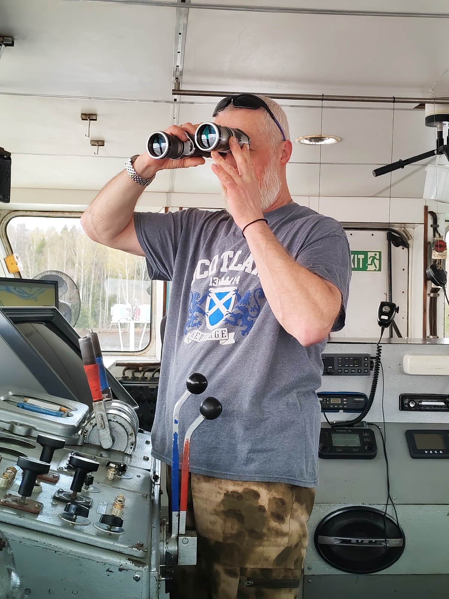 A man stands in a ship's bridge, looking through binoculars. He wears a grey t-shirt with a blue crest, camouflage pants, and a silver watch on his left wrist. Sunglasses rest on top of his head. The bridge contains various control panels, switches, levers, a microphone, radio equipment, and an exit sign with a green figure. A window shows trees and water outside. The location is Unknown, Unknown.
