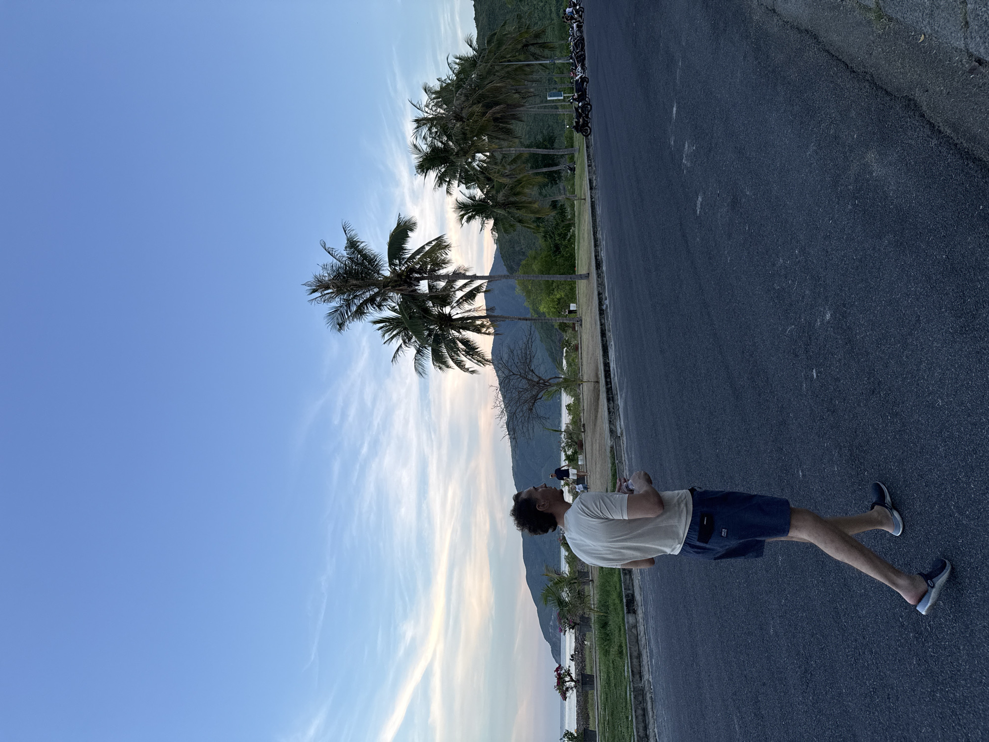 A man is standing on an asphalt road, looking up at the sky. He is wearing a light-colored t-shirt and dark shorts. The image is oriented vertically, with the man appearing to be walking into the frame. In the background, there are palm trees, mountains, and the ocean. The sky is a clear blue with some wispy clouds. The sun appears to be setting or rising, casting a warm glow on the scene. The overall mood is peaceful and relaxed. The location is identified as Nam Nha Trang Ward, Vietnam.