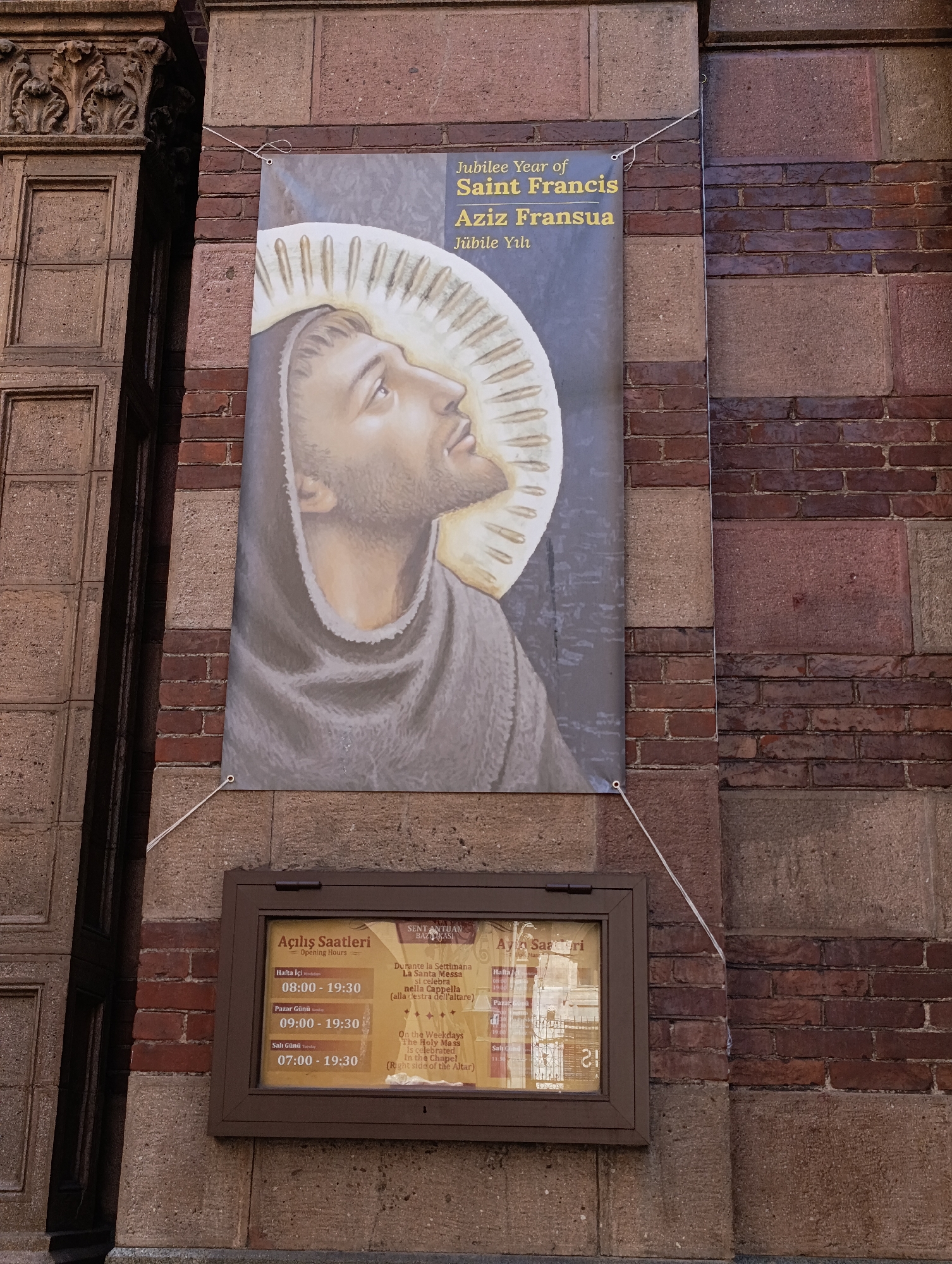 The image shows a banner depicting Saint Francis of Assisi with a halo, hanging on a brick wall. Below the banner, a framed notice board displays opening hours and information about mass times. The building appears to be a church or religious institution. The architectural style, with its stone and brickwork, suggests an older building. The banner and notice board are mounted on the exterior wall, which is made of alternating bands of stone and brick. The overall scene is one of a public building with religious significance. The information on the notice board is written in Turkish and Italian, and includes specific times for opening hours and mass. The banner's text is in English and Turkish. The context of the image points to Beyoğlu, Turkey, a district known for its historical and religious sites.