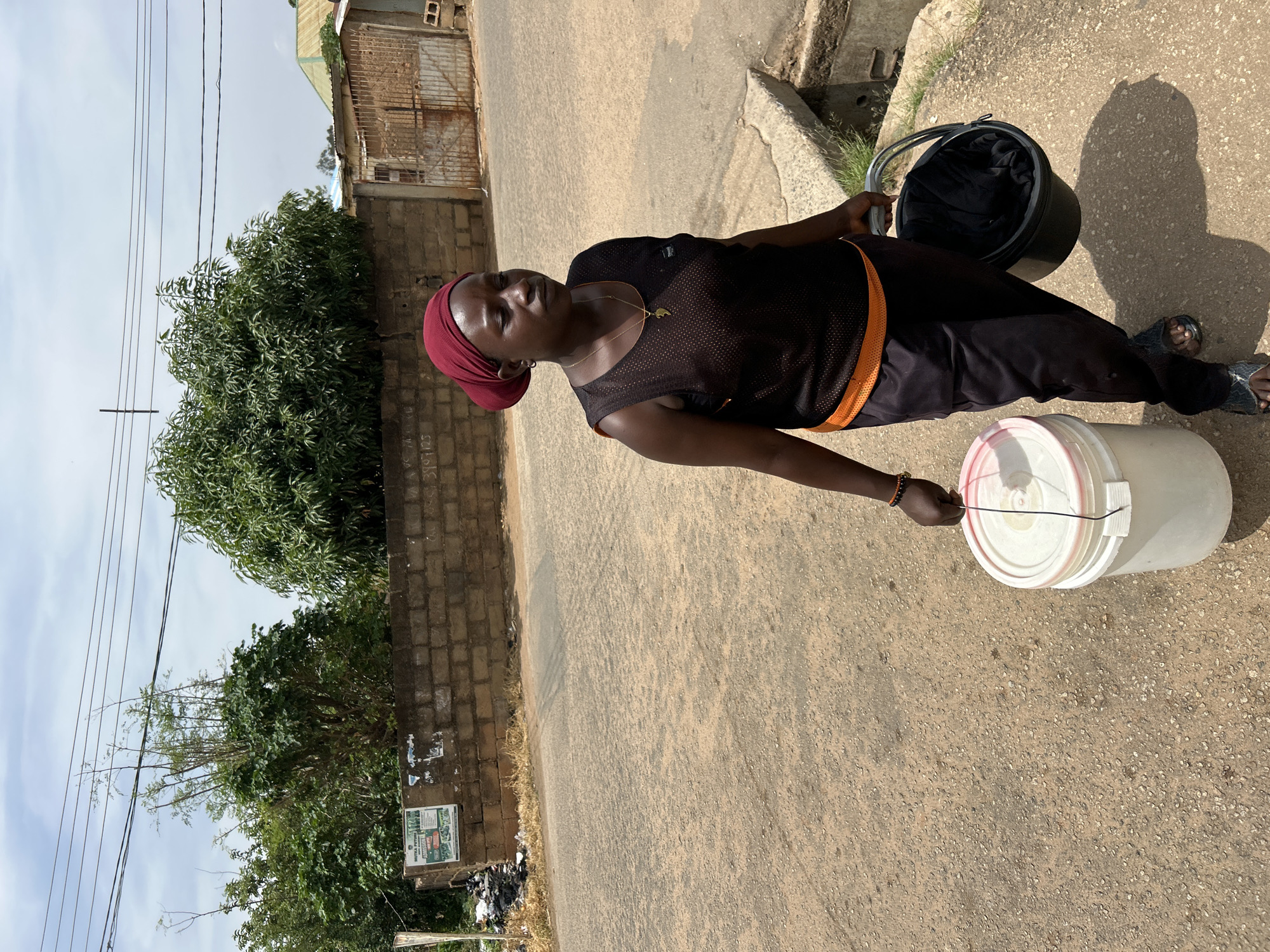 A Black woman, wearing a dark tank top over an orange band, black trousers, and sandals, walks along a paved road in Jos, Nigeria. She carries a large white bucket in her left hand and a smaller black bucket in her right hand. She is looking to her right. In the background, a brick wall separates the road from a row of trees and buildings. The sky is cloudy.