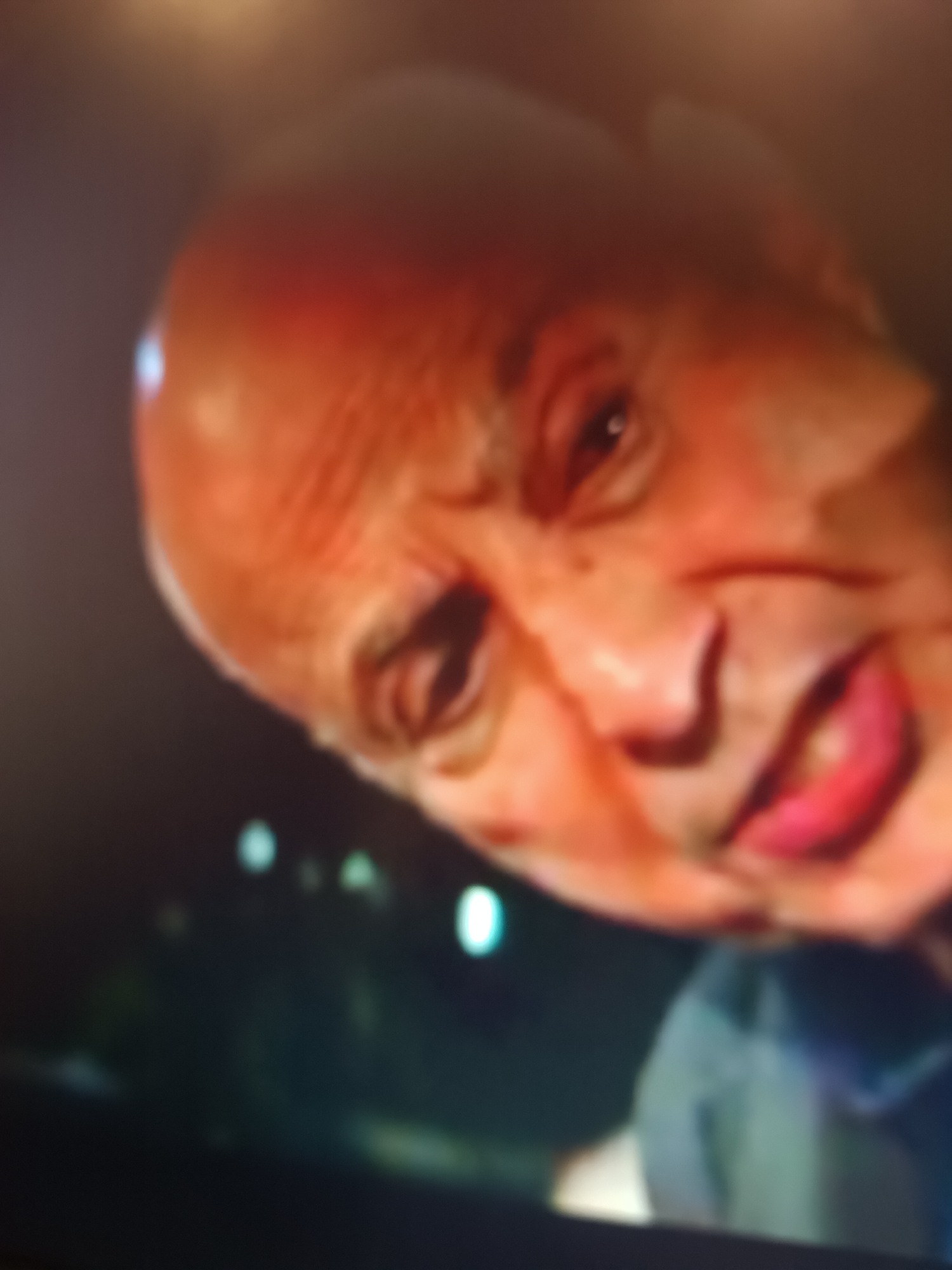 The image presents a close-up, angled portrait of an older, bald man. His face, illuminated with a reddish tone, is the central subject. Pronounced wrinkles are visible on his forehead and around his eyes, suggesting an advanced age. His lips are a striking, vibrant red. He has dark eyes and appears to be looking directly towards the viewer or slightly off-camera, with a somewhat ambiguous, possibly neutral or subtly contemplative expression.

The background is dark and significantly out of focus, featuring several blurred points of light, indicative of bokeh. This suggests the photograph was taken in a dimly lit environment, possibly at night, with artificial light sources in the distance. No specific objects or additional subjects are discernible in the blurred background. The lighting on the man's face is somewhat strong, highlighting his features. No activity is in progress other than the man's direct gaze. There is no visible text in the image. While the location context is Pchelarovo, Bulgaria, no visual cues within the image itself confirm this.