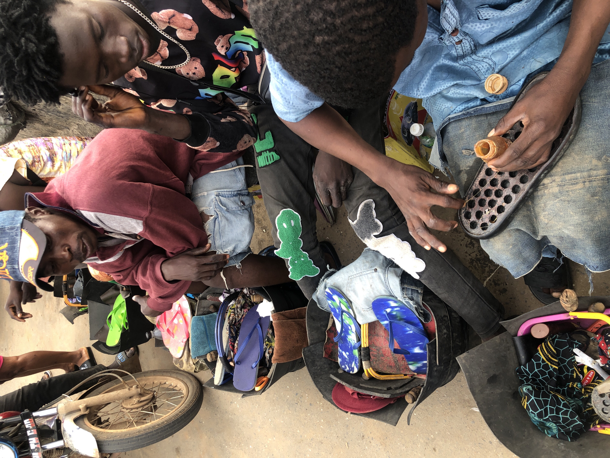 The photograph captures an outdoor scene in Ikeja, Nigeria, focusing on a group of individuals engaged in repairing footwear. In the foreground, a person in a blue shirt is carefully examining a sole and a small bottle of adhesive, suggesting they are in the process of shoe repair. Beside them, another person wears dark pants adorned with green patches and a patterned shirt featuring teddy bears. To the left, a person wearing a maroon hoodie and a baseball cap is seated, with their hands resting on their lap. A motorcycle with a visible wheel and fender is parked on the left side of the frame. Various containers filled with flip-flops, towels, and other assorted items are scattered around the individuals, indicating a mobile repair or vending setup. The ground appears to be paved, characteristic of an urban environment.