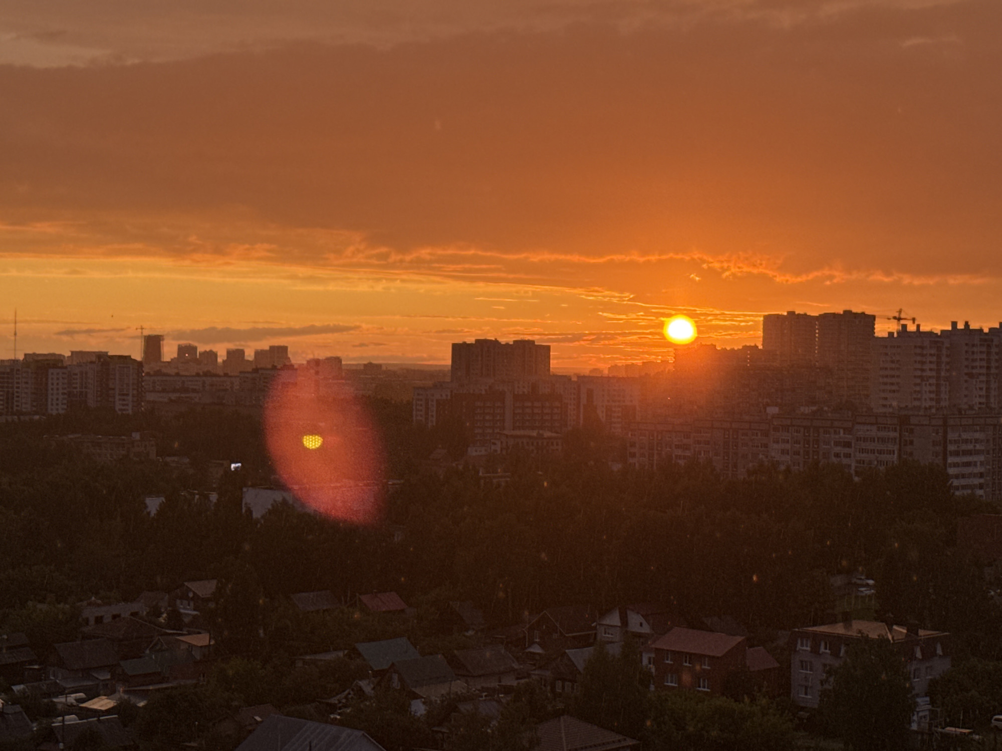 This is an aerial, wide-angle, medium-distance photograph of a sunset over a cityscape in Izhevsk, Russia. The sky is dominated by vibrant orange and yellow hues, with scattered clouds. The sun, a bright, blown-out white disk, is setting on the right side of the frame, casting a warm glow over the scene. In the foreground, a dense canopy of dark green trees obscures a residential area with houses and smaller buildings. Behind the trees, a line of multi-story apartment buildings stretches across the middle ground, with some taller structures visible in the distance. A construction crane can be seen among the buildings on the right. The image exhibits lens flare, with a prominent orange, heart-shaped artifact appearing on the left side of the frame. The overall atmosphere is one of a warm, atmospheric sunset over an urban environment.