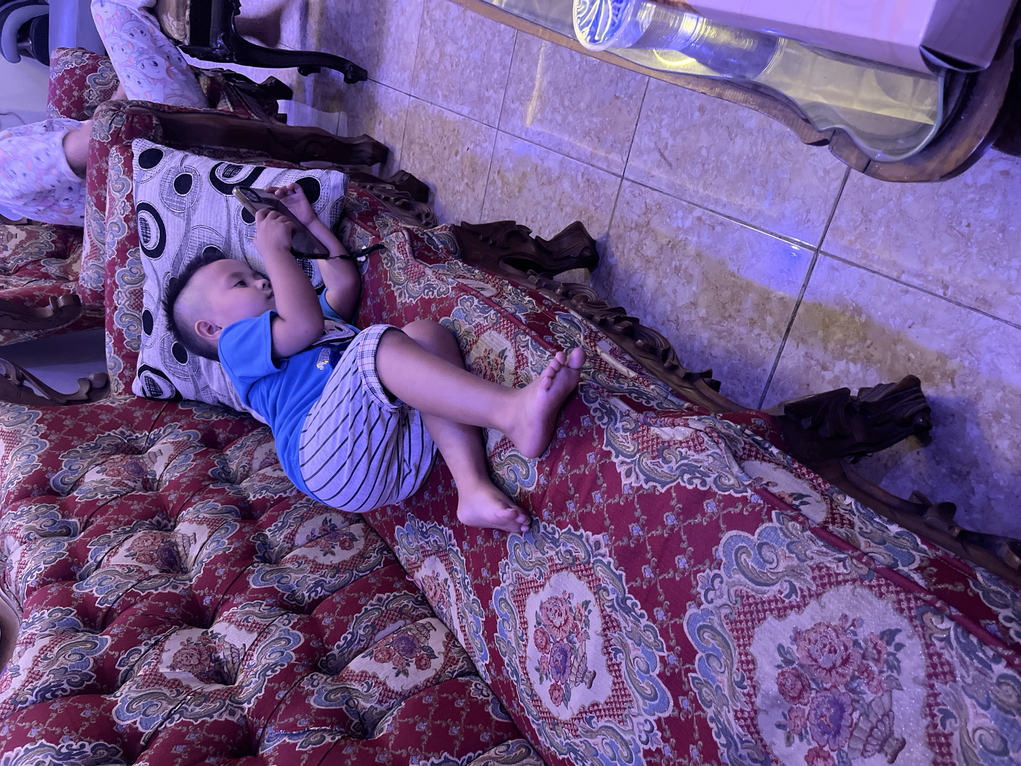 A young boy wearing a blue shirt and striped shorts lies on a red and white ornate sofa, holding a mobile phone. His hair is styled in a mohawk. The sofa is covered in a detailed floral pattern. In the background, another person wearing light-colored patterned clothing is visible, seated on a similar sofa. The floor is tiled with a light brown marble pattern. A glass-topped coffee table with a bottle of water and a box is positioned in the foreground. The lighting suggests an indoor setting, possibly a living room, with a purplish hue cast over the scene. The location is Jatisari, Indonesia.